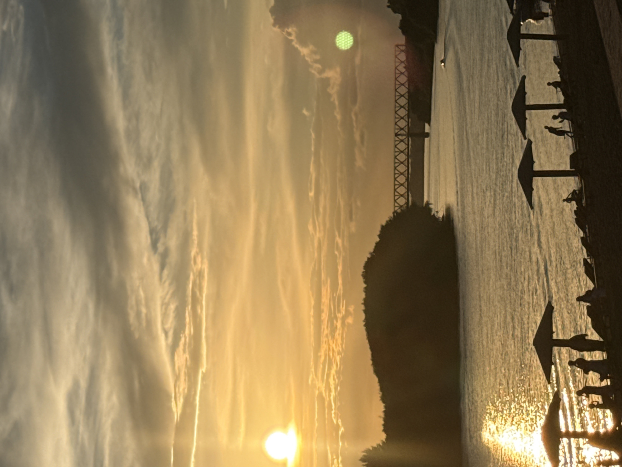 The image depicts a sunset scene at a riverfront, likely in the city of Pavlodar, Kazakhstan, given the presence of a distinctive railway bridge. The sun, a bright orb, is setting on the left side of the frame, casting a warm, golden glow across the sky and water. The sky is filled with a mixture of clouds, some thin and wispy, others more voluminous, catching the light in various shades of yellow, orange, and grey.
In the foreground, a sandy or pebbled area lines the riverbank. Several beach umbrellas are set up, casting dark shadows on the ground. Silhouetted figures of people can be seen scattered along the shore, some standing, others appearing to be sitting or relaxing. There are also lounge chairs visible. The river itself is calm, reflecting the bright colors of the sky and the setting sun, with shimmering highlights.
In the middle ground, a body of water, presumably the Irtysh River, flows towards the right of the frame. On the far bank, a tree-covered hill or island is visible, silhouetted against the bright sky. Spanning the river is a large railway bridge with a distinctive lattice structure, a prominent landmark of Pavlodar. A small boat or vessel is visible moving on the water in the distance, leaving a subtle wake. The overall atmosphere is one of tranquility and the end of a summer day.