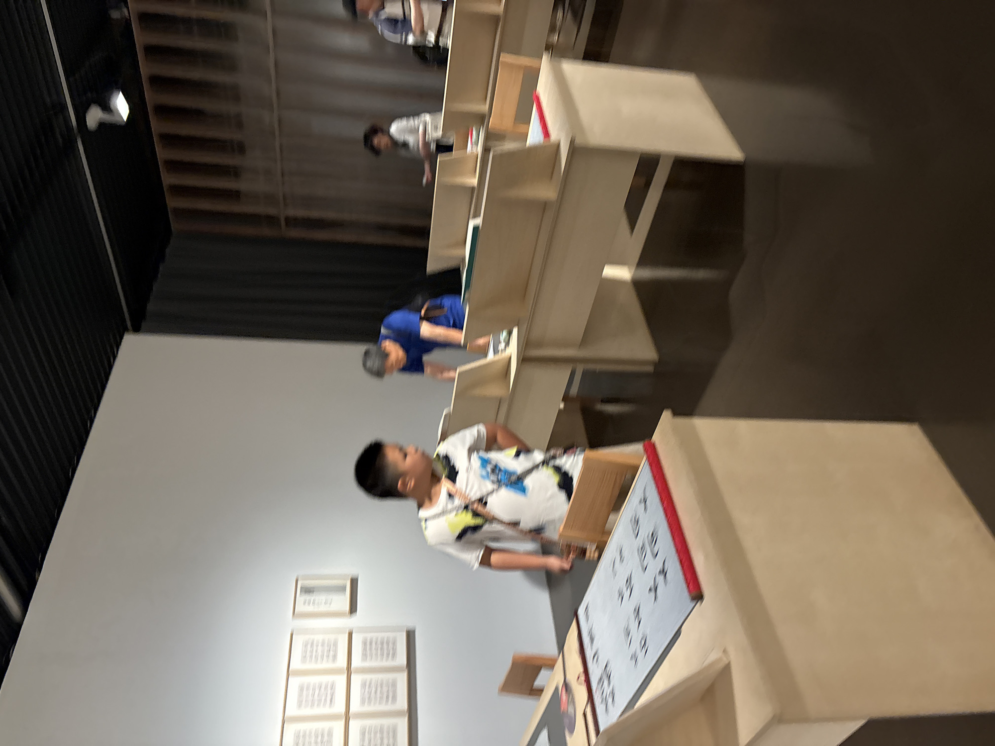 The image depicts an indoor art exhibition space, possibly in Pudong, China, with several people observing or interacting with the exhibits. In the foreground, a young boy wearing a white t-shirt with a colorful graphic print stands near a display table. The table holds a large, flat piece of fabric with rows of text in what appears to be an East Asian script, bordered by a red cylindrical object. To the left of the boy, a wooden chair is visible.
Further back in the scene, a man in a blue shirt is leaning over a similar display table, suggesting active engagement with the exhibit. Behind him, other individuals are present, including a woman in a white top and a man in a striped shirt, all appearing to be visitors at the exhibition. The space features light-colored walls and a dark ceiling with exposed lighting fixtures. Display structures made of light-colored wood are arranged in rows, each with partitions to hold exhibit items. The flooring is a dark, reflective surface. On the wall in the background, a grid of framed artworks or documents is displayed. The overall impression is that of a contemporary art gallery or museum setting.