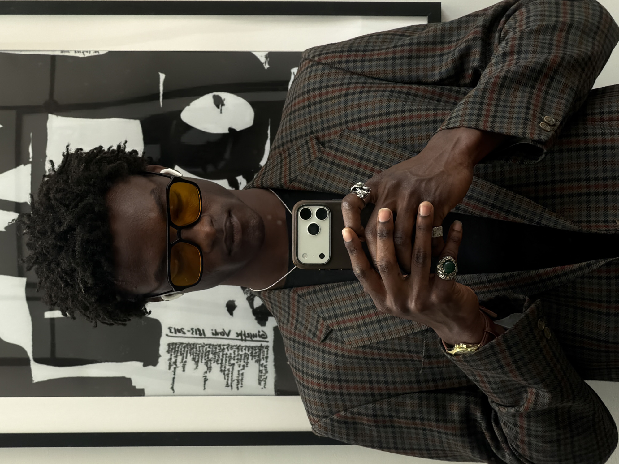 A medium shot depicts a dark-skinned person wearing a plaid blazer and yellow-tinted glasses, holding a smartphone to take a picture. The person has curly dark hair and is adorned with rings on their fingers. A silver necklace is visible around their neck.
Behind the person is a framed piece of art featuring bold black and white abstract designs and text.
The image does not contain enough information to determine if the location is in the city of London, United Kingdom.