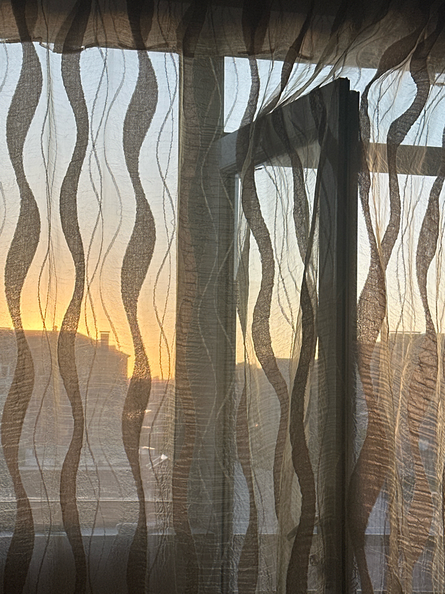 The image shows a close-up view of a sheer curtain with a wavy, vertical pattern. The curtain is partially covering a window, through which a sunset can be seen. The light of the setting sun casts an orange glow on the scene, illuminating the buildings and the sky outside. The curtain's translucent fabric distorts the view of the outside, creating a soft-focus effect. The image is taken from inside a room, looking out. The style of the buildings visible through the curtain suggests an urban environment, possibly Gyumri, Armenia, known for its historic architecture and the prominent presence of sunset views from many vantage points. There are no people or animals visible in the image, and no specific actions are taking place. The focus is on the interplay of light, fabric, and the urban landscape at dusk.