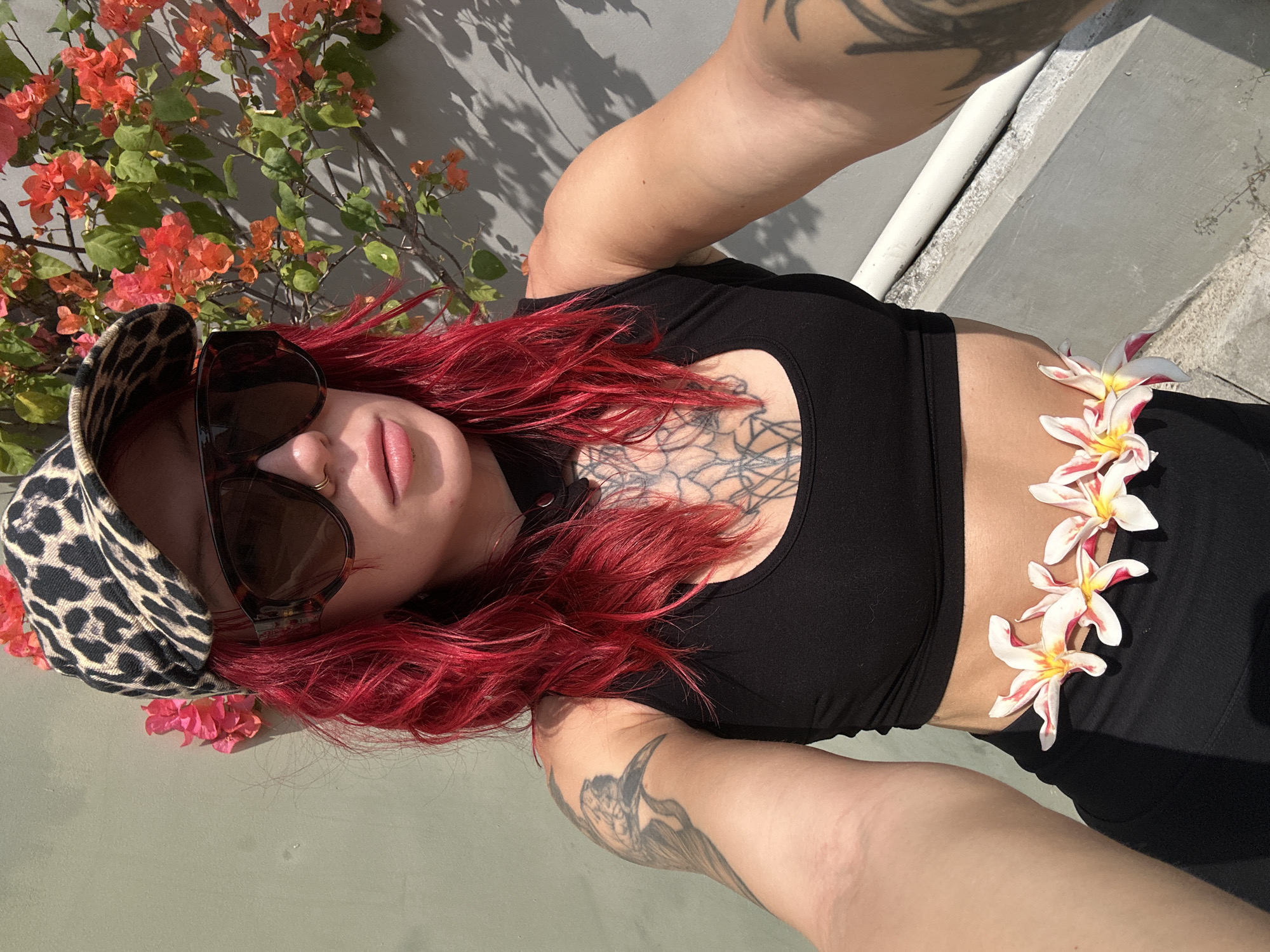 A woman with bright red hair and a leopard-print baseball cap is pictured from the chest up, taking a selfie outdoors. She is wearing large, dark sunglasses, a black crop top, and black leggings. A lei made of white and pink flowers is draped around her waist. Behind her, a grey wall is visible, with a vibrant bougainvillea plant in bloom. The scene appears to be set in a sunny, outdoor location, possibly a balcony or patio. The location is identified as North Kuta, Indonesia, suggesting a tropical environment.