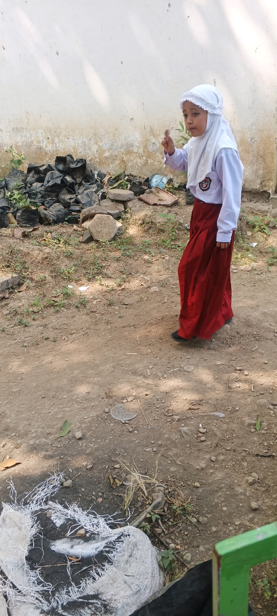A young girl, wearing a white hijab and a school uniform consisting of a white long-sleeved shirt and a red skirt, stands in what appears to be an outdoor area. She is looking to her left with her right hand raised, index finger extended, as if pointing or making a statement. Behind her, a light-colored wall shows signs of age and weathering. In the foreground and mid-ground, the ground is dirt and gravel, with scattered debris including black plastic bags filled with what looks like soil or compost, stones, and some dry vegetation. In the lower left corner, a torn white bag with dark contents is visible, and a green metal object is present in the lower right. The environment suggests an unkempt outdoor space, possibly on the grounds of a school or a residential area in Penanggungan, Indonesia. The lighting indicates it is daytime, with sunlight casting shadows on the ground.