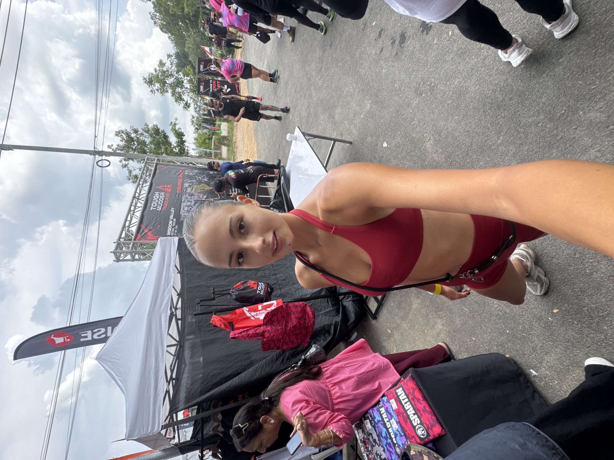Here is a description of the image:
The image appears to be a selfie taken outdoors at an event, likely a race or competition, in บ้านสำนักตะแบก, Thailand. The person taking the selfie is a young woman wearing a red sports bra and shorts. She has fair skin and her hair is pulled back. She is smiling slightly at the camera.
In the background, there is a crowded area with many people walking around. There is a banner that says "TOUGH MUDDER THAILAND, PATTAYA, THAILAND, WISDOM VALLEY" indicating the event and location. There are also some tents and tables set up. One tent has a white canopy and a flag with a red logo.
Another woman is bending over a table in front of the tent, looking at something, possibly a product or item for sale. She is wearing a pink top and has her hair down. There are various items on the table. The ground is paved and there are trees and power lines visible in the distance.