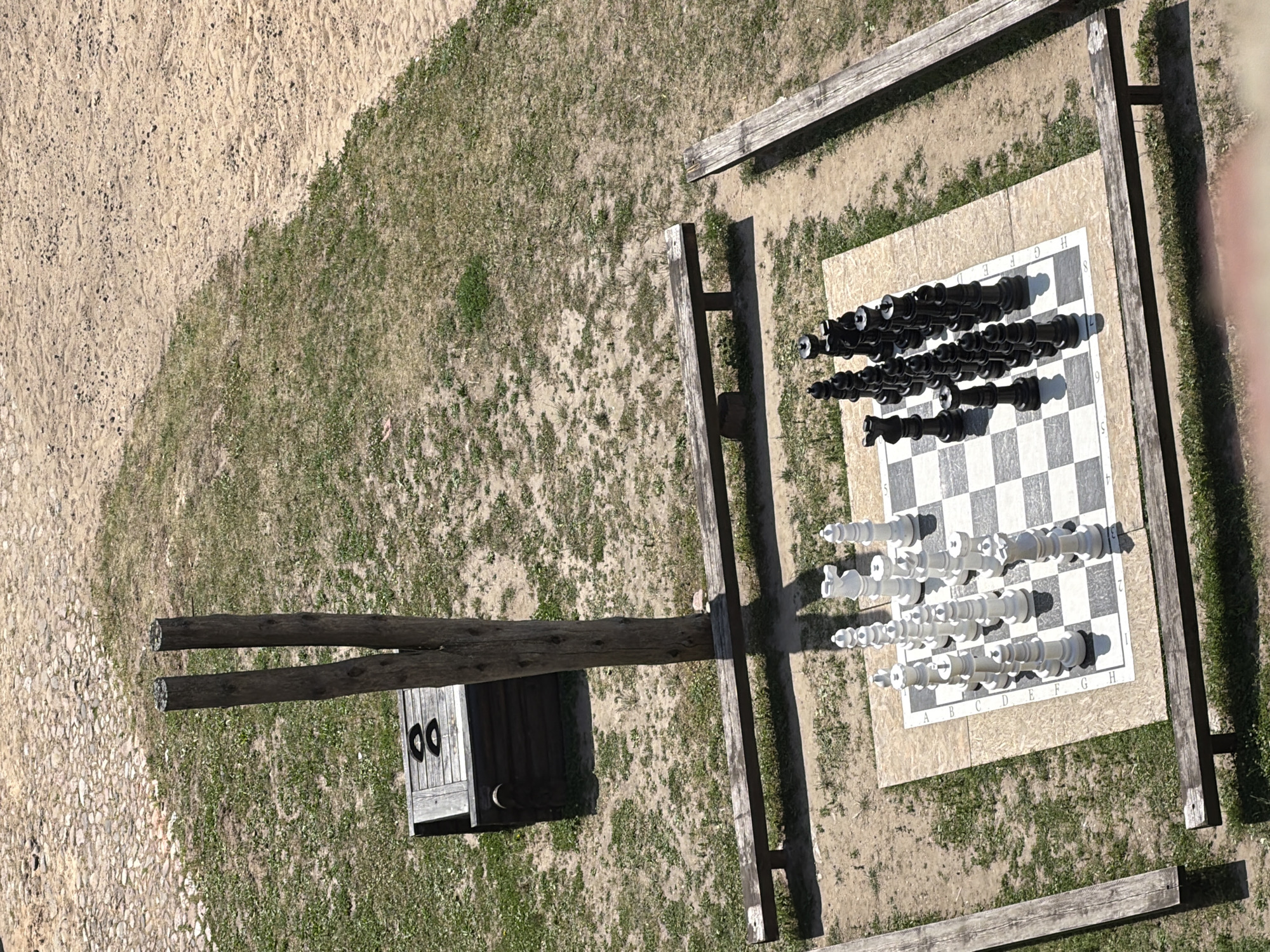 An outdoor giant chessboard is set up on a dirt and grass area, surrounded by wooden benches. The chessboard has white and black pieces arranged on it, suggesting a game in progress. Behind the chessboard, two tall wooden posts stand vertically. To the left of the posts is a dark wooden chest with two circular indentations on its lid. The scene is observed from a high angle, looking down on the chessboard and its surroundings. The ground is a mix of dry grass and sandy soil.