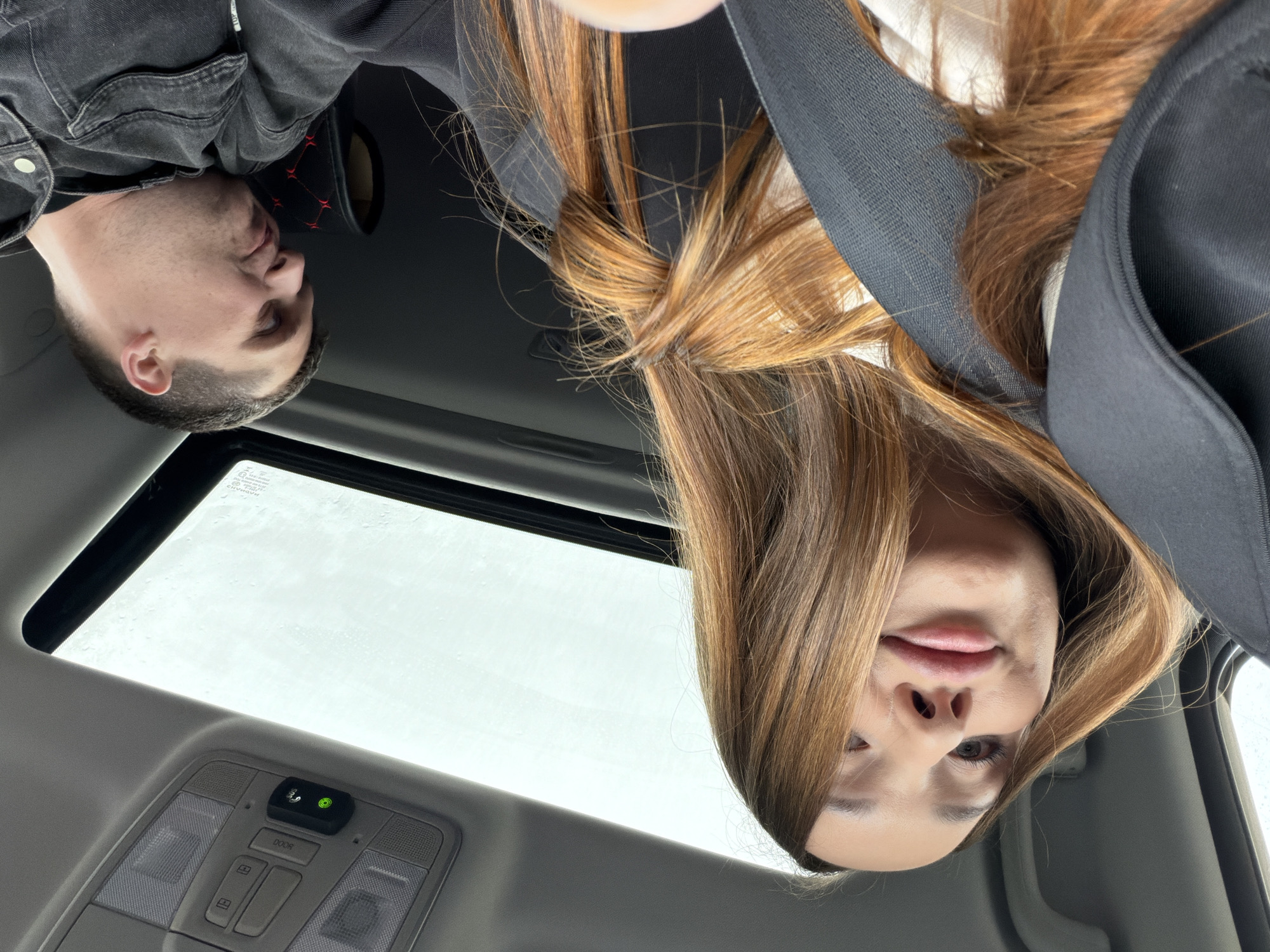 From a low-angle perspective inside a vehicle, the photo shows two individuals and the vehicle's interior. One individual, a man, is on the left wearing a black denim jacket. The other, a woman, is on the right with long brown hair and a black seatbelt across her chest. Above them is an open sunroof, revealing a white sky. In the foreground is the car's ceiling with light controls. There is no information in the photo that indicates that the location is Chaikovsky, Russia.