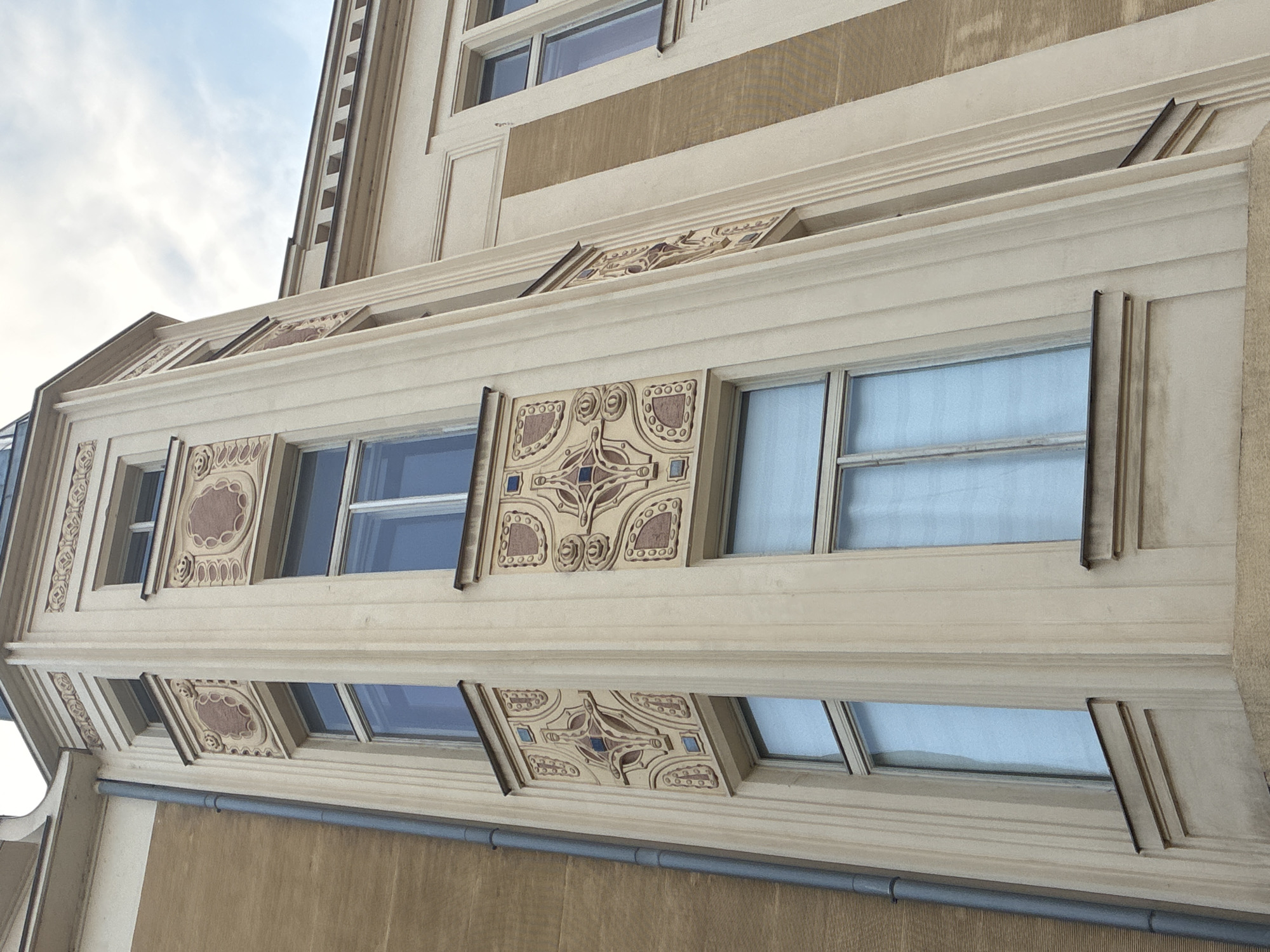 The image shows a close-up, low-angle view of the facade of an Art Nouveau building located in Prague, Czechia. The building features a corner tower with decorative relief panels and tall, narrow windows. The facade is adorned with ornamental elements in shades of cream, ochre, and terracotta, including geometric patterns and stylized floral motifs. A grey downspout runs vertically along the left side of the building. The sky is visible at the top of the frame, with light clouds.