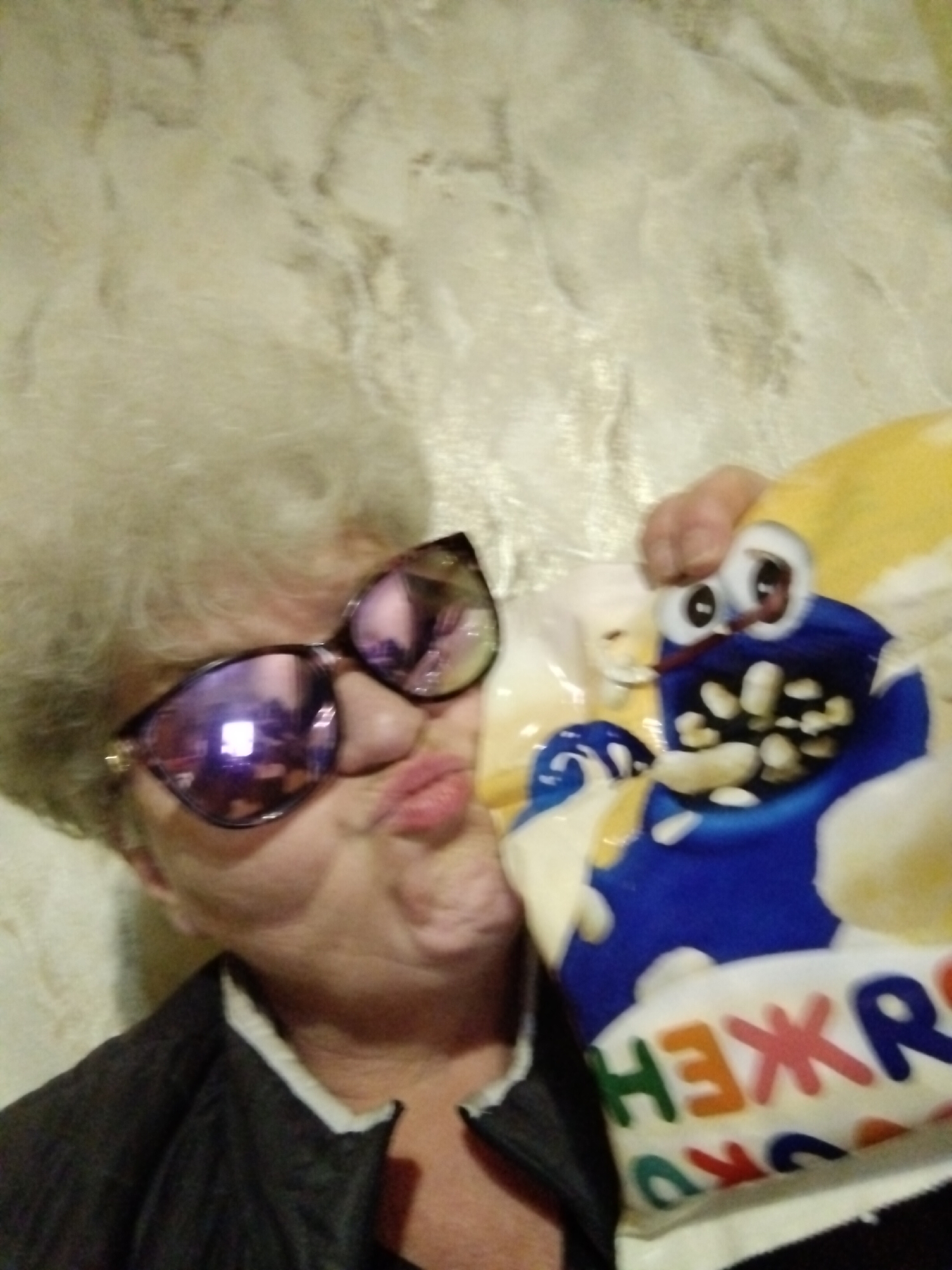 This close-up selfie captures a person, likely a woman, with voluminous grey hair, donning oversized, reflective sunglasses with purple hues. She is making a puckered "kissing" face, conveying a playful and lighthearted mood. Her left hand is visible, holding a brightly colored, inflated bag or pillow.
The bag features a cartoon character with a blue body, a yellow top section, large white eyes with black pupils, and an open mouth filled with irregular white shapes that resemble popcorn or snack pieces. Partially visible text on the bag is in Cyrillic, with distinct letters like "НЕЖР" and "ОКО", which, when combined with other less clear letters, spell out "СНЕЖОК" (Snezhok).
The individual is wearing a dark jacket, possibly black, with a white collar visible underneath. The background consists of a light-colored wall with a subtle, textured pattern, possibly mimicking marble or stucco, indicating an indoor setting. Reflections in the sunglasses show indoor lighting. The location context provided is Horishni Plavni, Ukraine.