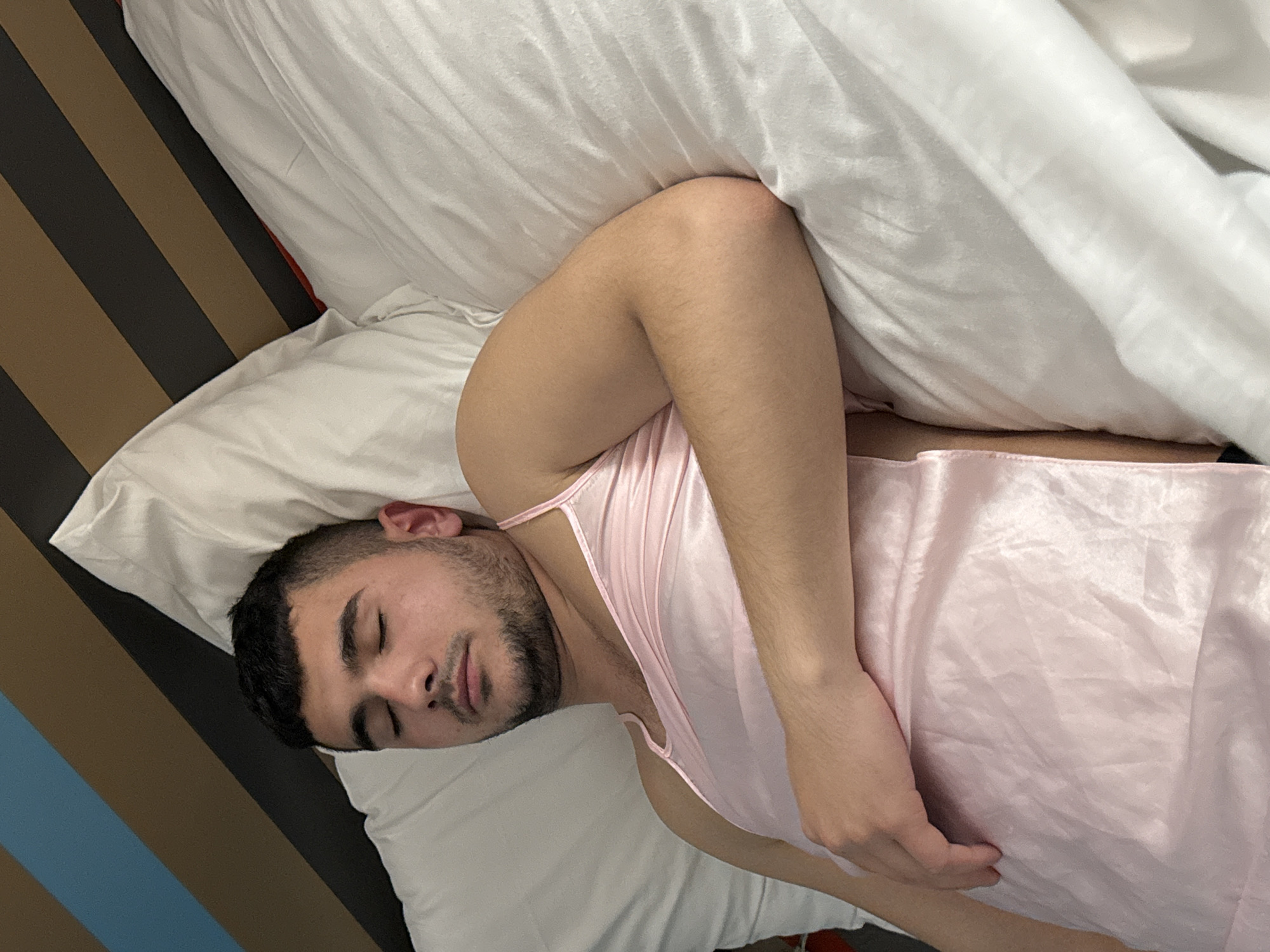 A man is sleeping in a bed. He is lying on his back, with his head resting on a white pillow. He is wearing a pink satin nightgown. His eyes are closed and his face is relaxed. The bed is covered with white sheets and pillows. The headboard of the bed has vertical brown, black, and blue stripes. The image does not provide any information to indicate the location is Antony, France.