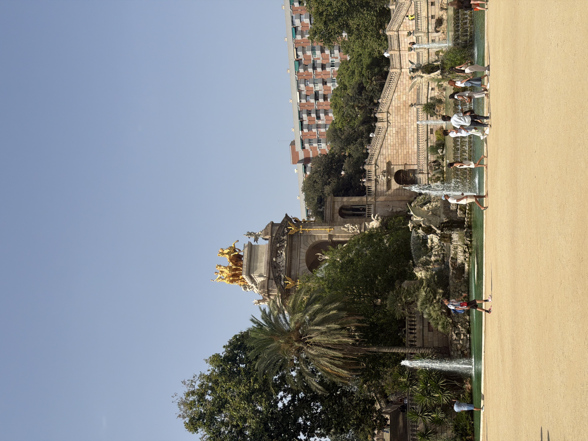 The image captures a sunny day at Parc de la Ciutadella in Barcelona, Spain. In the background, a grand fountain structure with golden statues of horses and chariots is visible. Adjacent to the fountain, a large stone staircase leads up to a building with multiple balconies, likely an apartment complex, painted in red brick. Lush green trees surround the fountain and the staircase, contributing to the park-like atmosphere. In the foreground, a sandy area is occupied by several people walking. A man in a white shirt and shorts stands near a fountain with a camera, taking a picture. Another man, wearing a white shirt, black pants, and carrying a backpack, walks across the sand. A group of individuals, mostly women, are seen walking together by the edge of the fountain's water. The water itself is a vibrant turquoise, with several fountains active and creating a misty spray. The sky is clear and blue, indicating good weather.