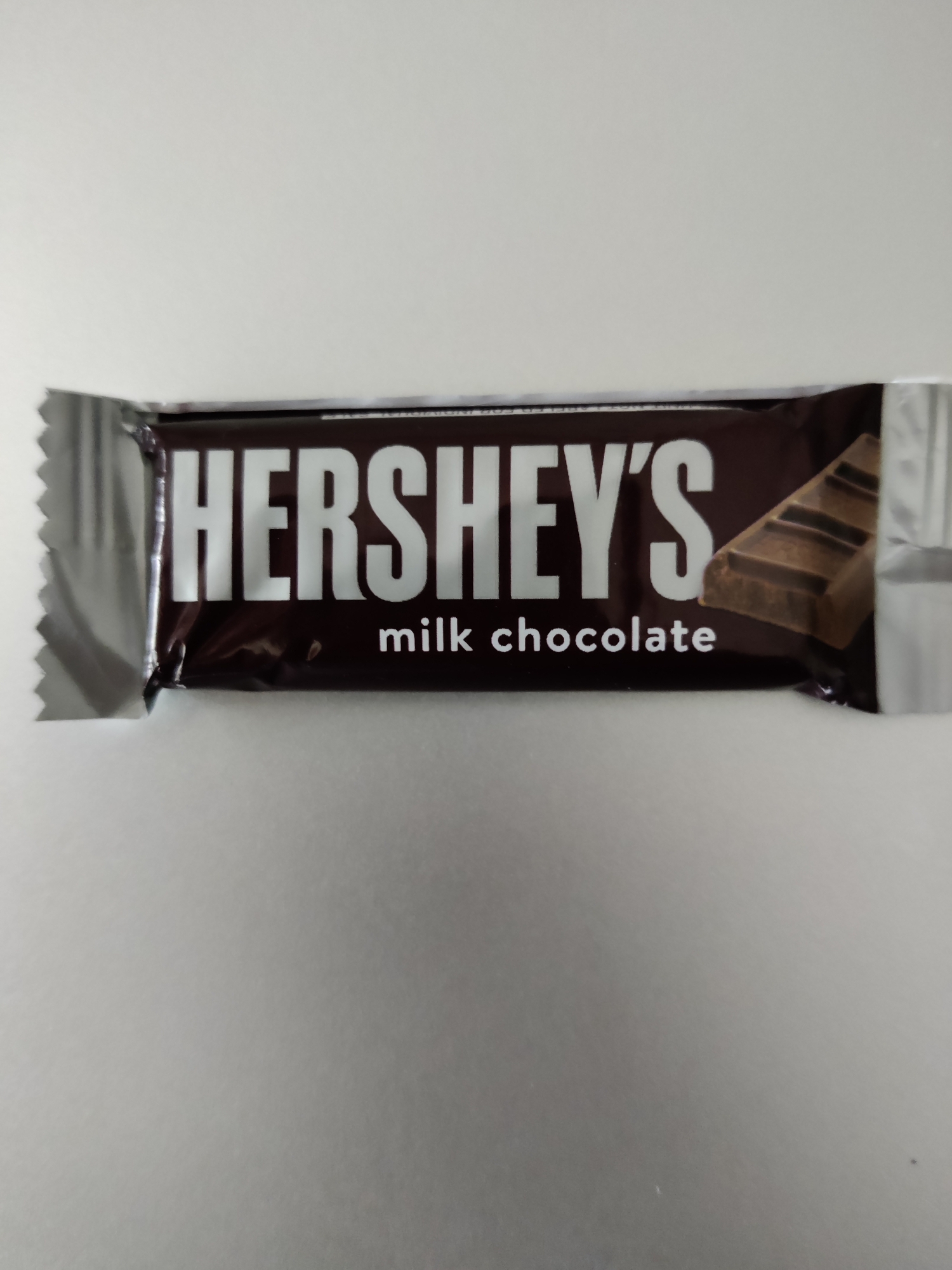 A Hershey's milk chocolate bar wrapper is centered on a light gray, textured surface. The wrapper is primarily dark brown with white lettering. The word "HERSHEY'S" is prominently displayed in large, bold, white capital letters. Below it, in smaller white lettering, are the words "milk chocolate." To the right of the lettering, a segmented piece of milk chocolate is visible through a transparent section of the wrapper, showing distinct squares. The left edge of the wrapper is silver and has a jagged, torn appearance. The background is out of focus and appears to be a plain wall or surface. There are no people or other objects in the image. The location is not discernible from the image.