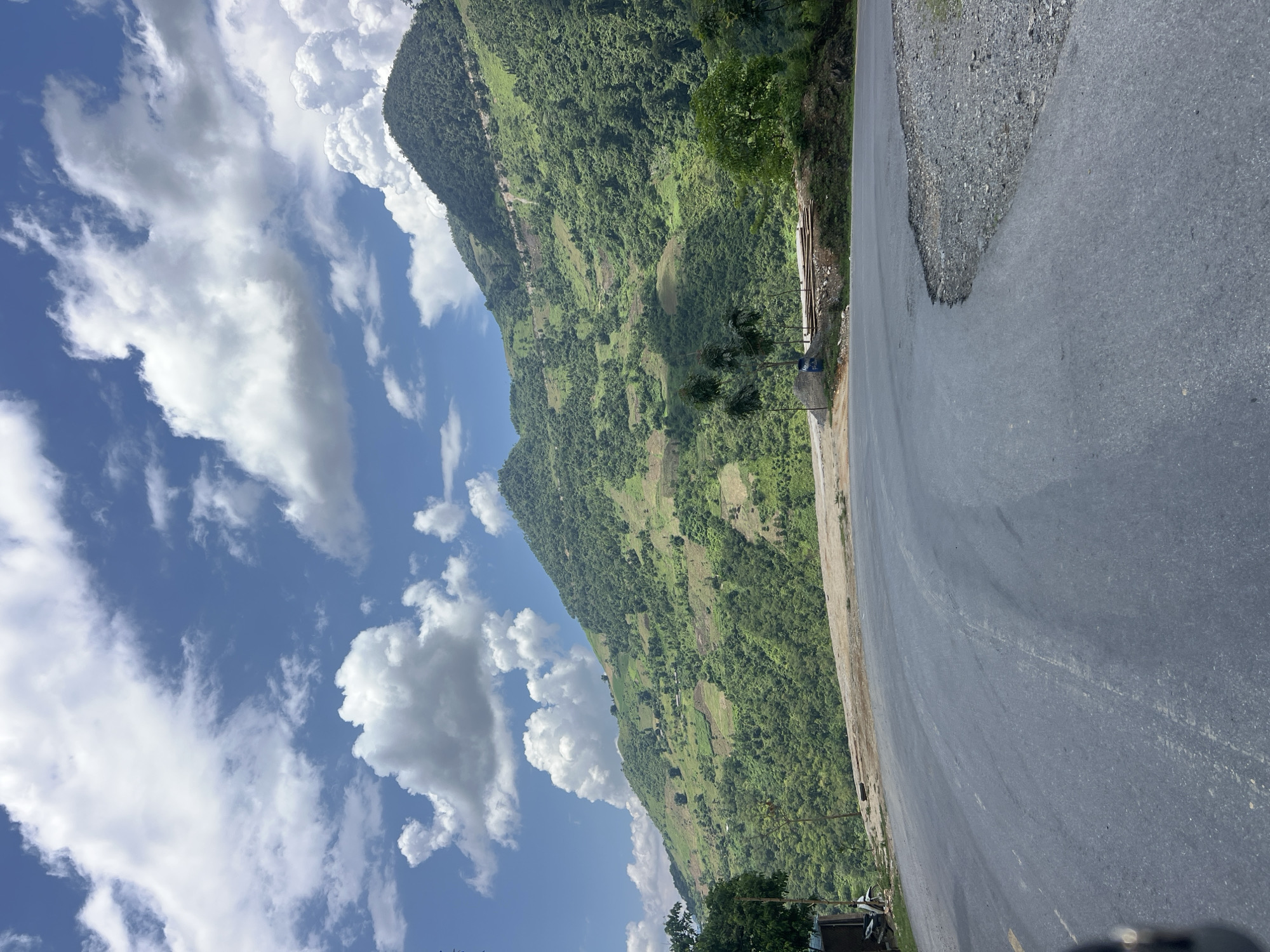 This is a landscape photograph taken in Minh Tân Commune, Vietnam. The image features a paved road curving into the foreground, with a steep, lush green mountainside rising in the background. The mountain is covered in dense vegetation, with patches of cultivated land visible on its slopes. Several trees dot the mountainside and the roadside. The sky above is a bright blue, dotted with fluffy white cumulus clouds. The road appears to be a secondary route, with gravel on the shoulder. The overall scene depicts a rural, mountainous area of Vietnam on a sunny day.