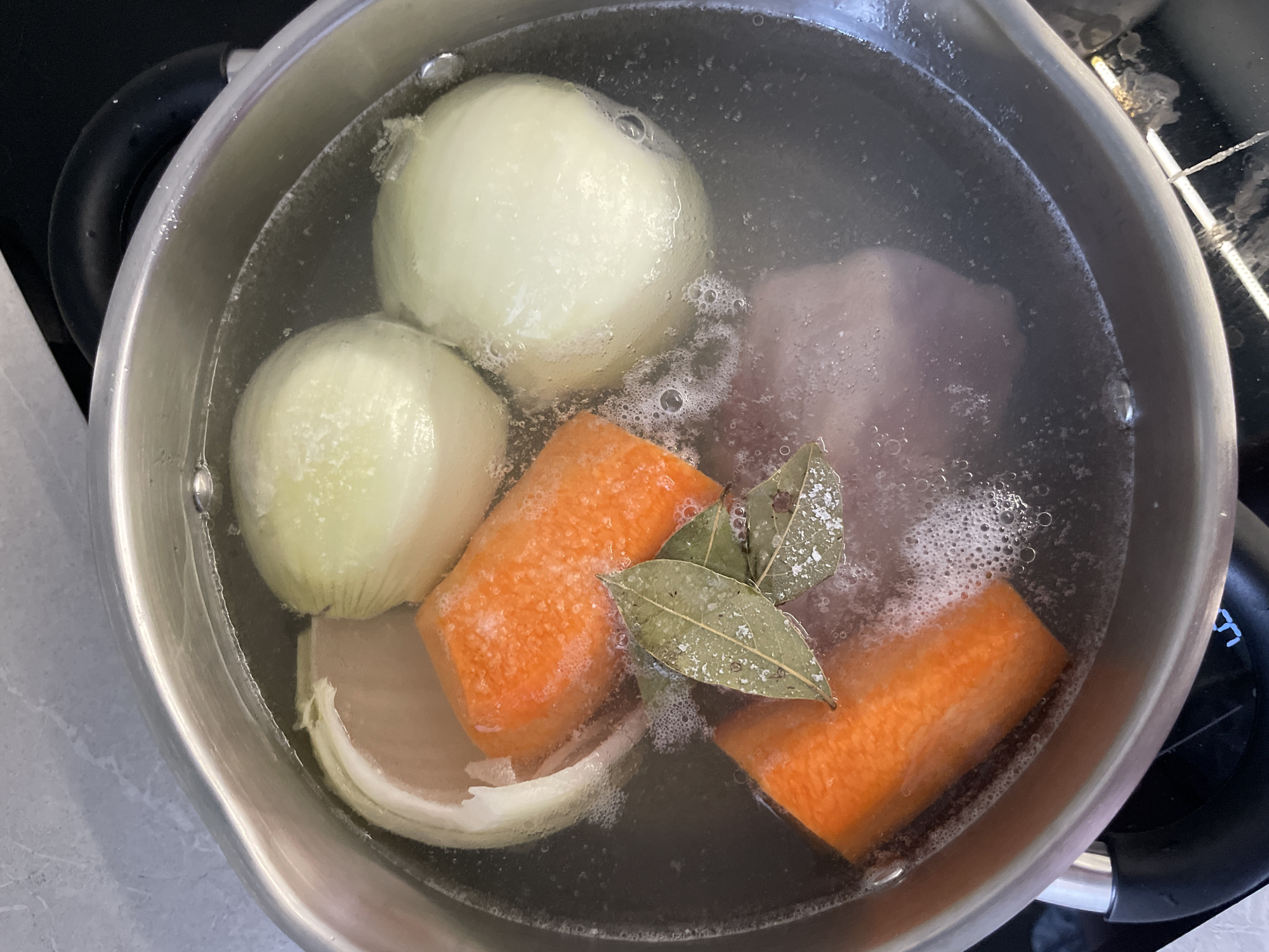 A top-down, eye-level shot shows a stainless steel pot filled with water and various ingredients. Two whole, peeled onions are visible along with a partial third onion, two large, peeled carrot chunks, and two bay leaves. The pot is on a black cooktop. The ingredients are submerged in water. There is no information available to determine whether the image was taken in Izhevsk, Russia, or elsewhere. No people are visible in the image.