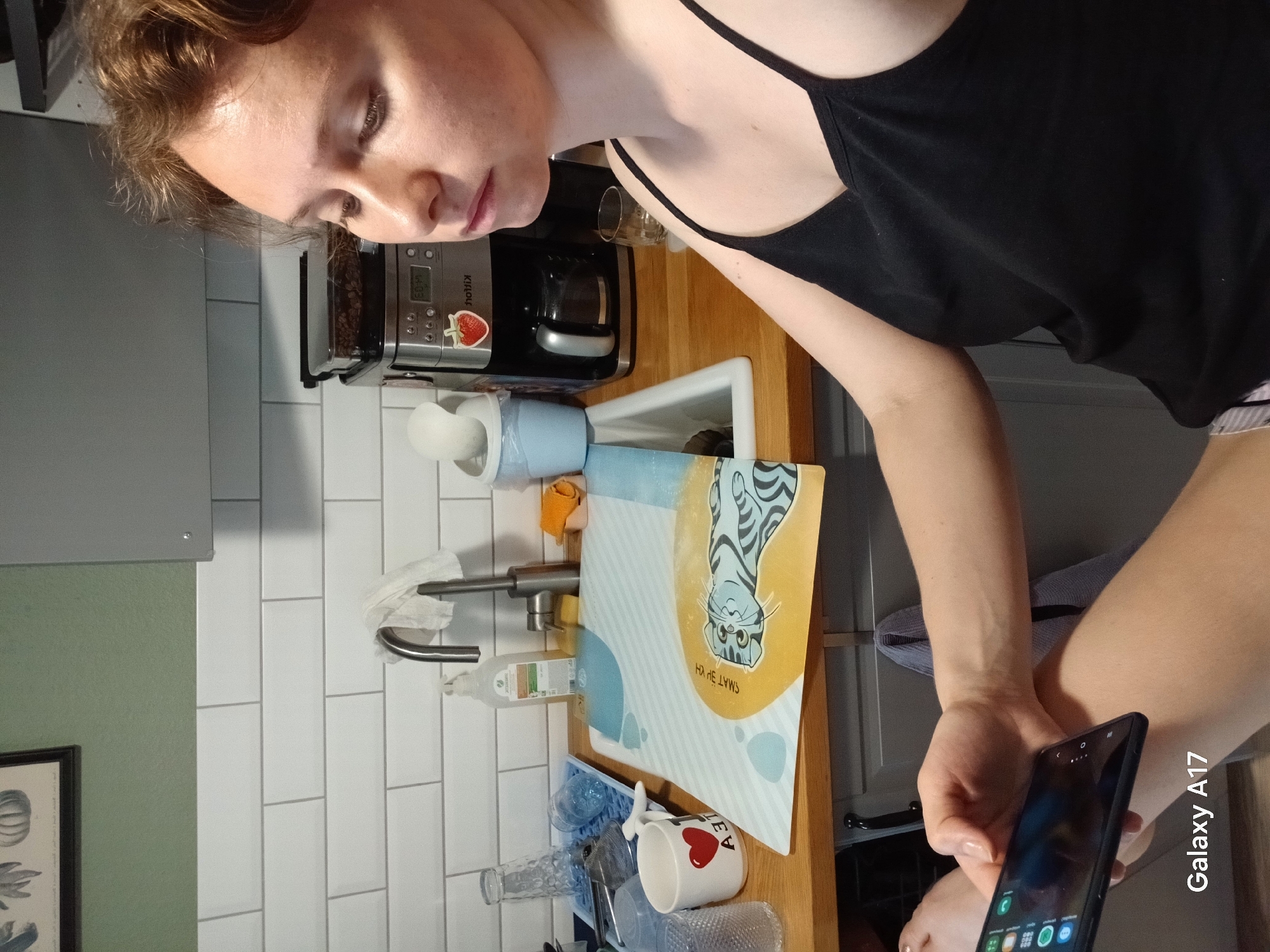 A woman in a black tank top is holding a black smartphone, labeled "Galaxy A17," in a kitchen. A coffee maker sits on the counter next to a sink with a dish rack holding various items including a mug with a heart, a patterned dish towel, and several glasses. The wall behind the sink is covered in white subway tile and a green painted section with a framed botanical print. The location in the city of Yekaterinburg, Russia is not discernible from the provided image.