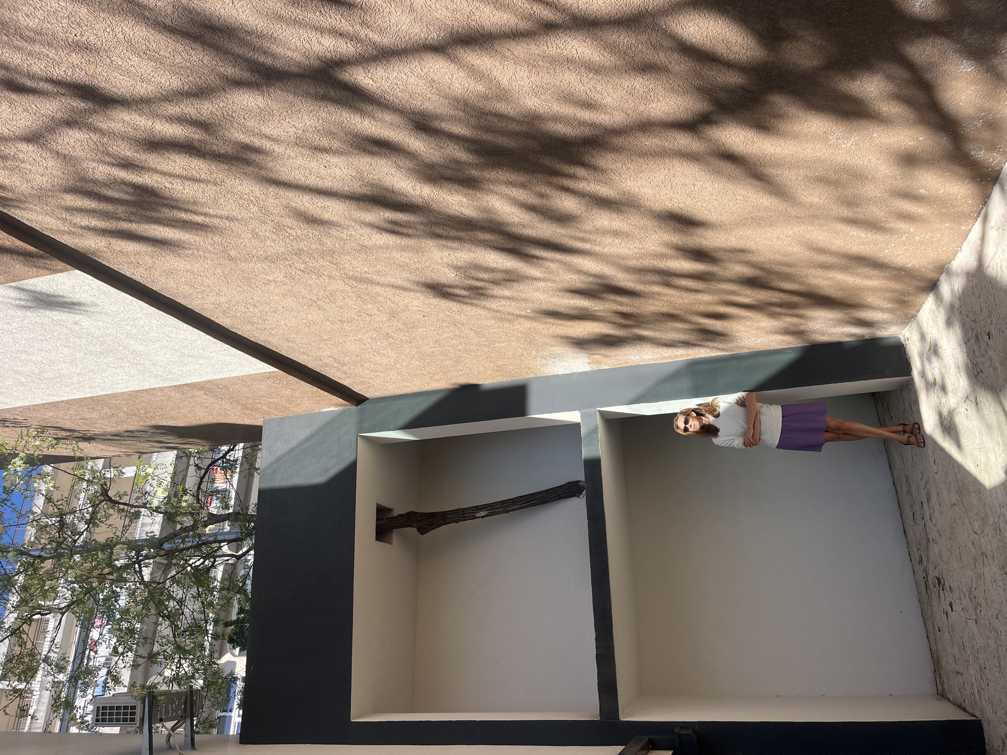 The image depicts a woman standing in an alcove. The woman is wearing sunglasses, a white top, a purple skirt, and brown sandals. She is positioned in the right alcove, facing forward. A tree branch is placed horizontally in the left alcove. The alcoves are part of a structure, located in Budva, Montenegro. In the background on the left is a building with many windows. The top of the image contains shadows from a tree on a beige surface. The angle of the photo is oriented at a slight upward angle, looking towards the ceiling.