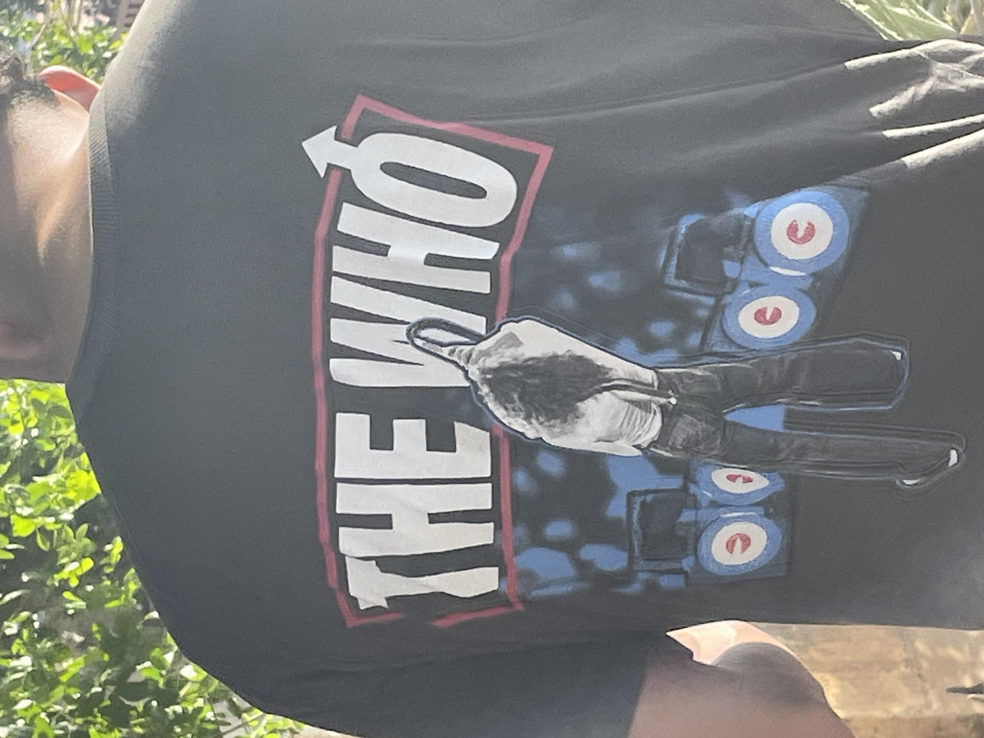 The rear view of a person wearing a black t-shirt with a graphic print of the band "The Who" is shown. The graphic features the band's name in large white letters with a red outline, and a symbol resembling Mars. Below the text, there is an illustration of a person with long hair, wearing jeans and suspenders, standing in front of two blue amplifiers with red and white circular logos. The scene is outdoors, with green foliage visible in the background. The lighting suggests it is daytime.