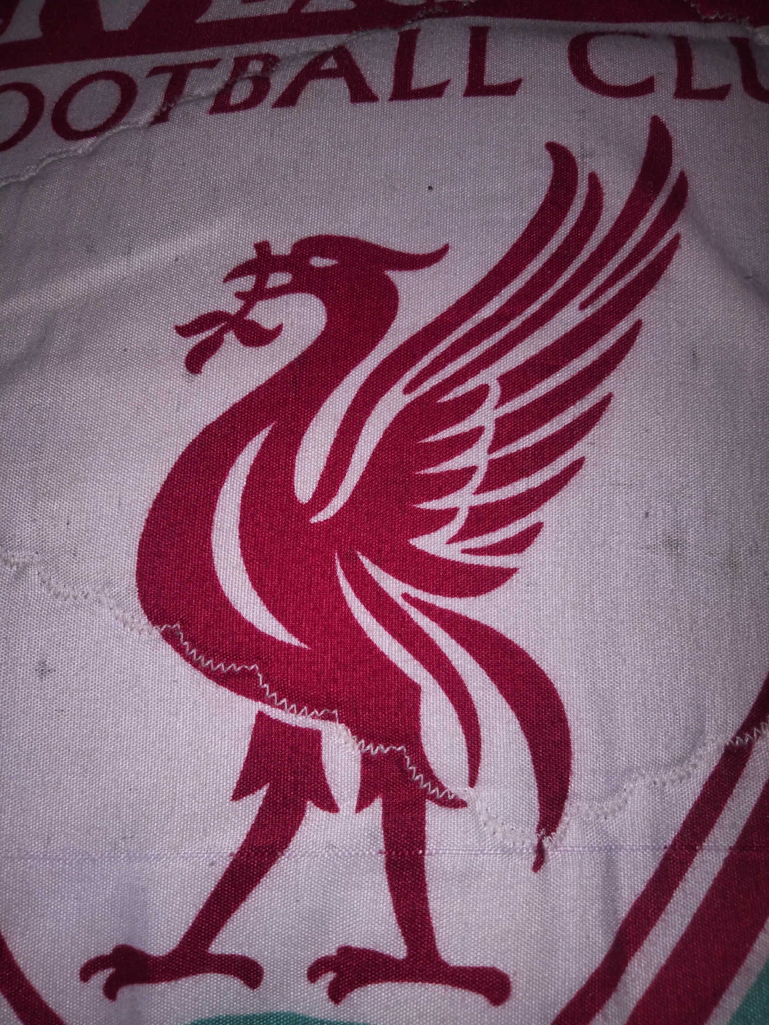 The image displays a close-up view of a fabric with a red Liverbird crest and partial text reading "OOTBALL CL". The fabric appears to be a part of a garment or textile item. The crest is detailed with red and white elements, and the text is also in red. There are visible stitches and some minor imperfections on the fabric. The image is an object study and does not depict a scene in Payagaboh, Indonesia, or any people or actions.