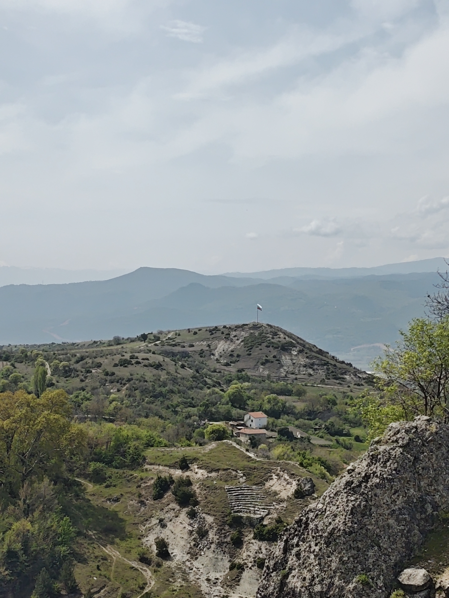 The image captures a wide panoramic view of a mountainous and hilly landscape under a bright, cloudy sky. In the foreground, large grey rock formations and green, leafy trees and shrubs frame the bottom right and left.
The midground features rolling hills covered in sparse green and brown vegetation, indicative of dry terrain. A small, white house with a reddish-brown roof is nestled among trees on one of these hills. To the left of the house, a series of terraced steps or seating areas are carved into the hillside. Further back, a prominent, conical hill rises, topped with a flagpole from which a flag with horizontal stripes of white, green, and red (from top to bottom) is flying, strongly suggesting the flag of Bulgaria. Dirt paths or roads snake across the hillsides.
In the background, multiple layers of distant mountains stretch across the horizon, appearing hazy and blue due to atmospheric perspective. The sky above is light blue with scattered white clouds, suggesting clear weather. The overall scene depicts a serene, vast natural environment, likely captured during daytime, possibly late morning or early afternoon. No visible text is present in the image.