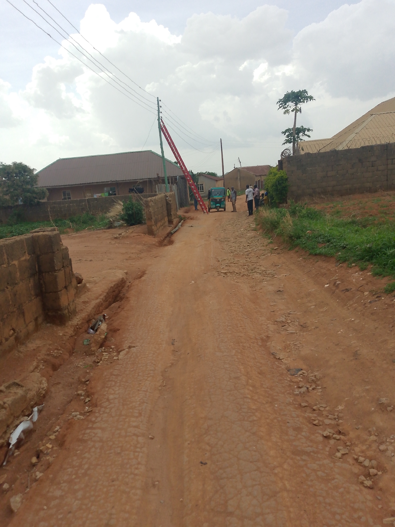 The image depicts a dirt road in Jos, Nigeria, with a red ladder leaning against a utility pole. The road is cracked and eroded with a small open drainage ditch to the left. Several people are gathered near a three-wheeled vehicle, interacting amongst themselves. In the background are single-story buildings and brick walls, typical of residential areas. Overhead are power lines stretching across the scene under a cloudy sky.
