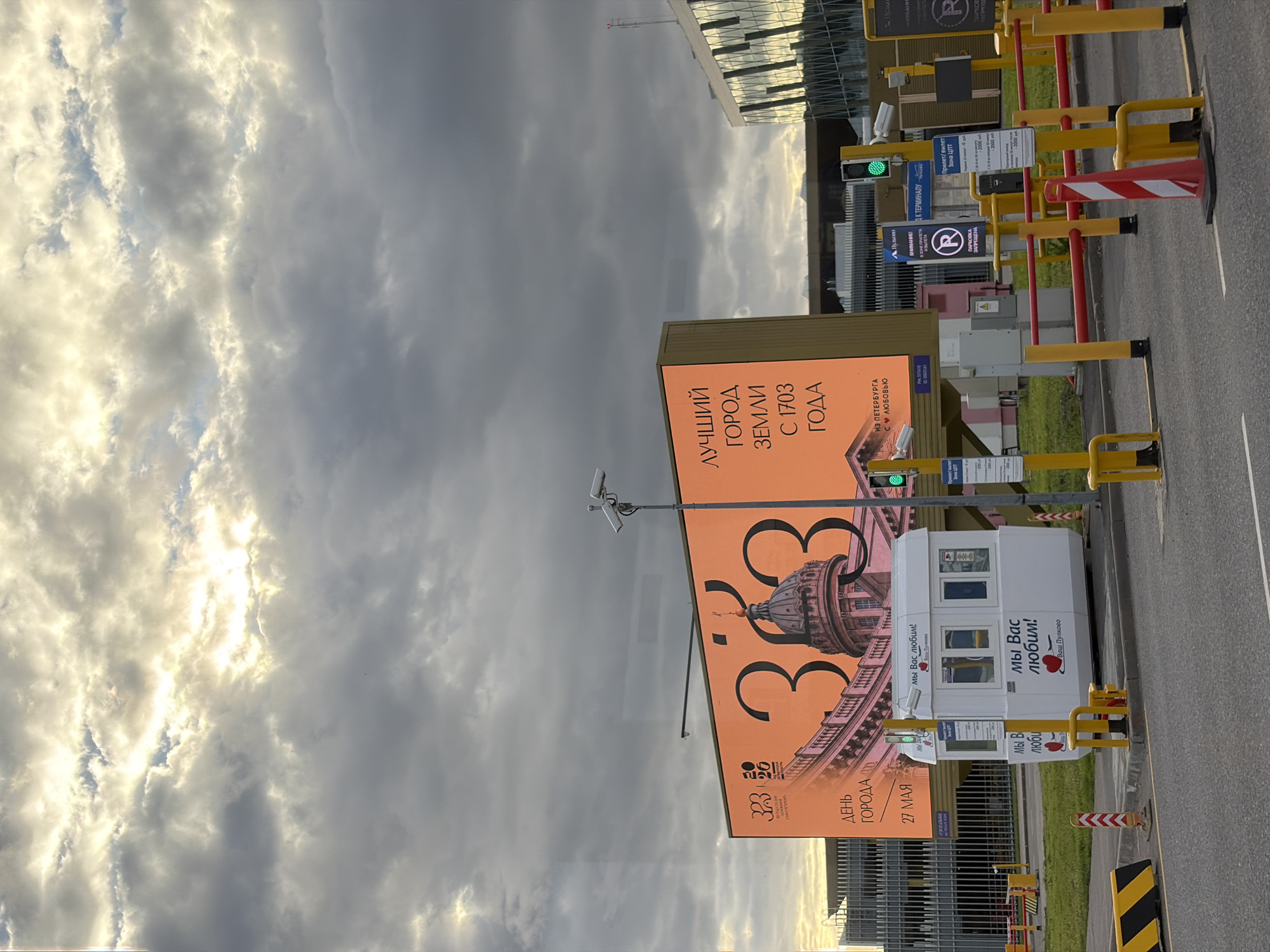 The image depicts a scene likely near Pulkovo Airport in Saint Petersburg, Russia. The sky is filled with dramatic clouds. A large advertisement displays "323 ДЕНЬ ГОРОДА 27 МАЯ" (323 Day of the City, May 27) along with the text "ЛУЧШИЙ ГОРОД ЗЕМЛИ С 1703 ГОДА" (Best City on Earth Since 1703), and the text "ИЗ ПЕТЕРБУРГА С ЛЮБОВЬЮ" (From Saint Petersburg With Love). There is a guard booth with the words "Мы Вас любим! Ваш Пулково" (We Love You! Your Pulkovo). Yellow and red safety barriers are present near signage for the arrival and departure zones, including parking information. The logo "R" is present. A traffic light is visible, showing green. The image gives the impression of a parking area leading to a terminal.