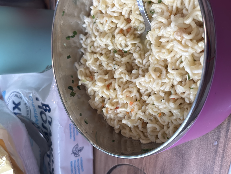 The image captures a close-up, slightly elevated view of a meal in Tatarsk, Russia. The primary subject is a circular insulated bowl with a bright pink exterior and a silver metallic rim, occupying the lower and central portion of the frame. The bowl contains a mass of cooked, light-yellow instant noodles, distinguished by their wavy, helical structure. Scattered throughout the noodles are small green fragments, consistent with chopped chives or similar herbs, and occasional minute orange fragments, likely diced vegetables. A silver spoon is partially submerged within the noodles on the right side of the bowl, indicating active consumption or preparation.
In the blurred background, to the upper left, a white plastic bag featuring Cyrillic text, including the word 'ВОДА' (water) and a large 'X', is visible. Beneath this bag, a smaller white package, potentially containing napkins or wipes, also displays Cyrillic text and an illustrative leaf pattern. To the upper right, a portion of a light blue or teal insulated cup or bottle is present. The objects rest upon a light brown surface, consistent with a wooden table or countertop. No individuals are visible in the scene.