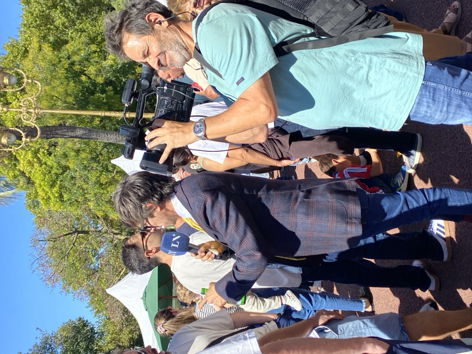 An outdoor scene captures a public event under a clear blue sky, likely within Plaza Francia or a similar park in Recoleta, Buenos Aires, Argentina, indicated by the distinctive ornate lamppost in the background. The ground is a reddish-brown gravel or crushed stone.
In the foreground and mid-ground, a crowd of people is gathered. Two central male figures are engaged in media coverage. On the left, a man in his 40s-50s, wearing a blue and red plaid blazer, yellow tie, blue jeans, and white sneakers, holds a blue microphone with a white "LV+" logo in his left hand. His right index finger is extended, pointing forward, as if asking a question or making a point. To his right, a man in his 50s-60s, with graying curly hair and a beard, is holding a black professional video camera (Sony brand) to his right shoulder, looking through the viewfinder. He wears a light blue short-sleeved t-shirt, blue jeans, a silver watch on his left wrist, and has an earbud in his left ear, with a black backpack strap visible over his right shoulder. He exhibits a slight smile.
In the background, lush green trees are visible, along with multiple white tents featuring green undersides, suggesting a fair or market event. Various individuals are scattered throughout the scene; notable figures include a person holding a baby in white attire with patterned shoes behind the reporter, a man in a white t-shirt and sunglasses, and a child in a red and white striped shirt bending down near the ground between the two central media figures.