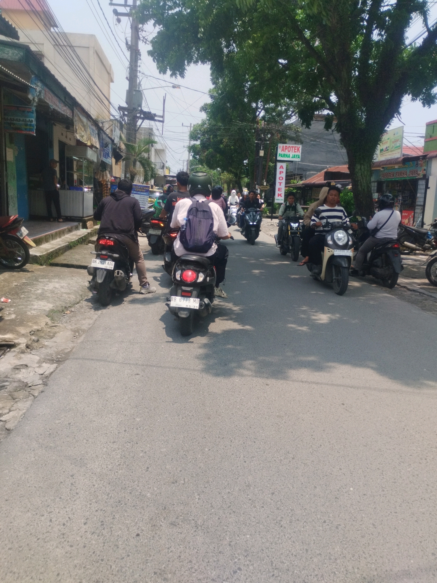The image depicts a street scene in the city of Medan, Indonesia, with a group of motorcyclists navigating the road. Several buildings line the left side of the street, including shops and what appears to be a laundry service. A prominent sign for "APOTEK PARNA JAYA" is visible on the right side of the street, indicating a pharmacy. The road surface is asphalt, with some signs of wear and tear.
In the foreground, a motorcyclist wearing a black jacket and light brown pants is riding a black scooter. Behind them, another motorcyclist is on a black scooter with a blue backpack. Several other motorcycles and riders are scattered throughout the scene, some in motion and others stationary. The environment is characterized by trees providing shade, overhead power lines, and a bright, sunny sky. The overall atmosphere suggests a typical day in a bustling urban environment.