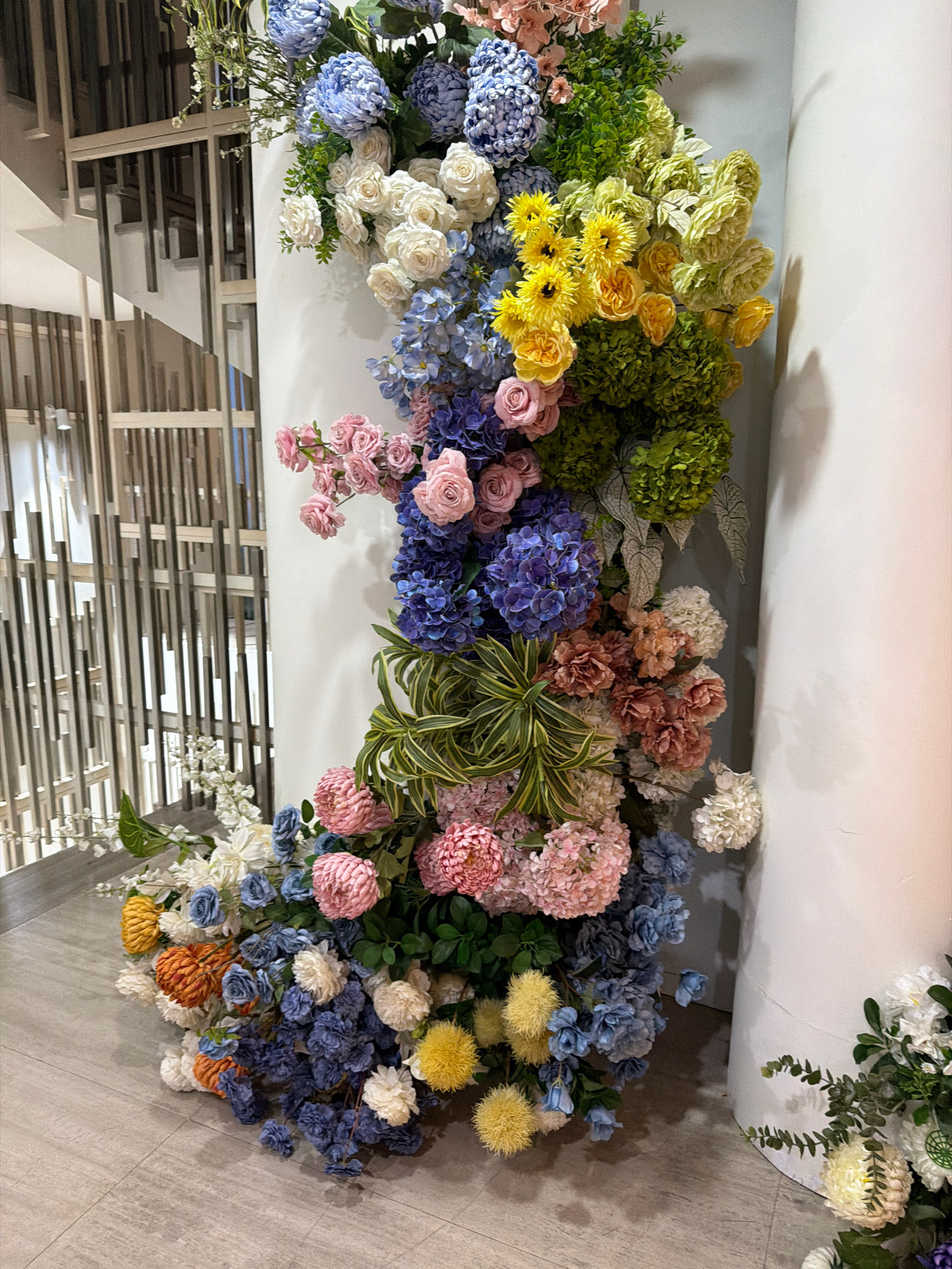 A vibrant floral arrangement cascades down a cylindrical white pillar in an indoor setting. The flowers are a mix of blues, pinks, whites, yellows, and greens, with various textures and shapes including hydrangeas, roses, and clusters of smaller blossoms. The arrangement starts at the top of the pillar and trails down to the floor, where it spreads out. Behind the pillar, a staircase with horizontal wooden slats is visible, suggesting an interior location like a hotel lobby or a commercial building. The floor is tiled in a light gray wood-grain pattern. There are no people or animals visible in the image. The lighting suggests an indoor environment with some ambient light creating shadows on the pillar. The overall impression is one of elaborate floral decoration.