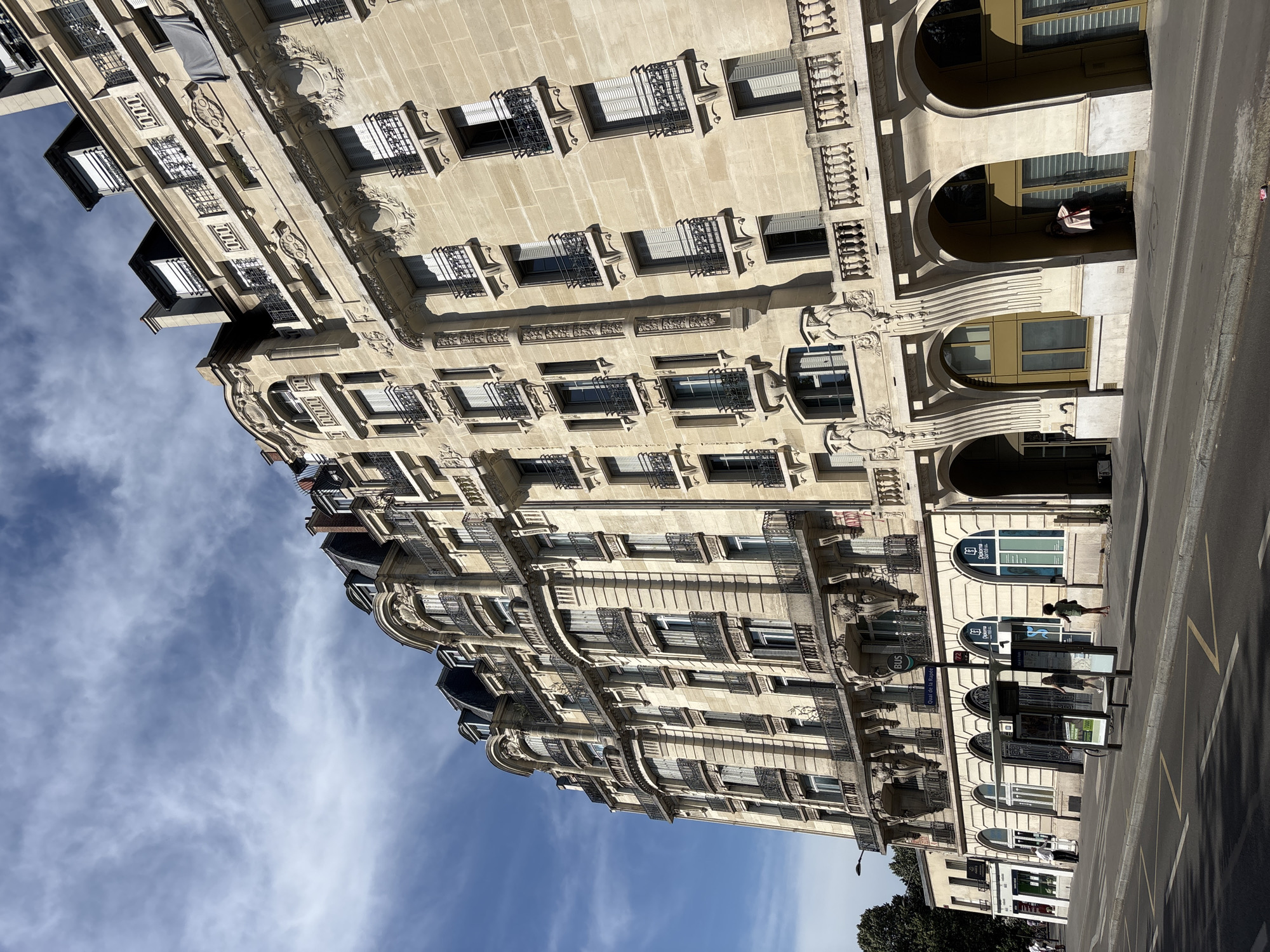 An eye-level, wide shot shows a tall, ornate building in Paris, France, under a bright blue sky with wispy white clouds. The building, made of light-colored stone, has multiple stories featuring numerous windows with black wrought-iron balconies. Elaborate architectural details, including decorative carvings and arched entrances on the ground floor, are visible. A street runs along the foreground, with a sidewalk and a bus stop. The bus stop displays advertisements, and a bus route sign indicates "Quai de la Rapée". A few people are present on the sidewalk: one person stands near the bus stop, another walks away from it, and a third individual is partially visible in an arched entryway of the building. The scene captures a typical urban streetscape in Paris during the daytime.