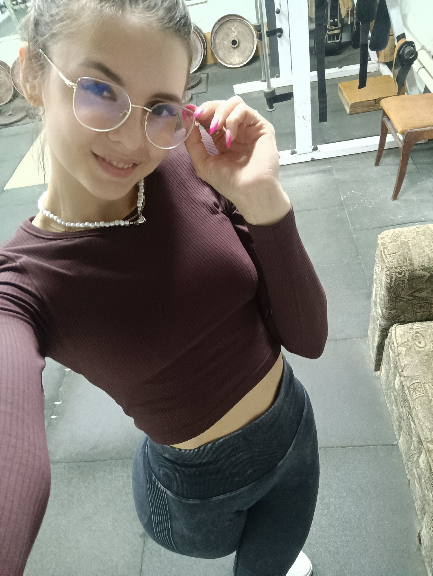 A woman is in what appears to be a gym or exercise area, based on the equipment in the background. She is wearing a long-sleeved maroon crop top, gray leggings, and white shoes. She is also wearing gold-rimmed glasses and a pearl necklace. She is holding the glasses with her right hand and smiling. In the background, there are weights, a weightlifting machine, and a stool. The floor appears to be gray tile.