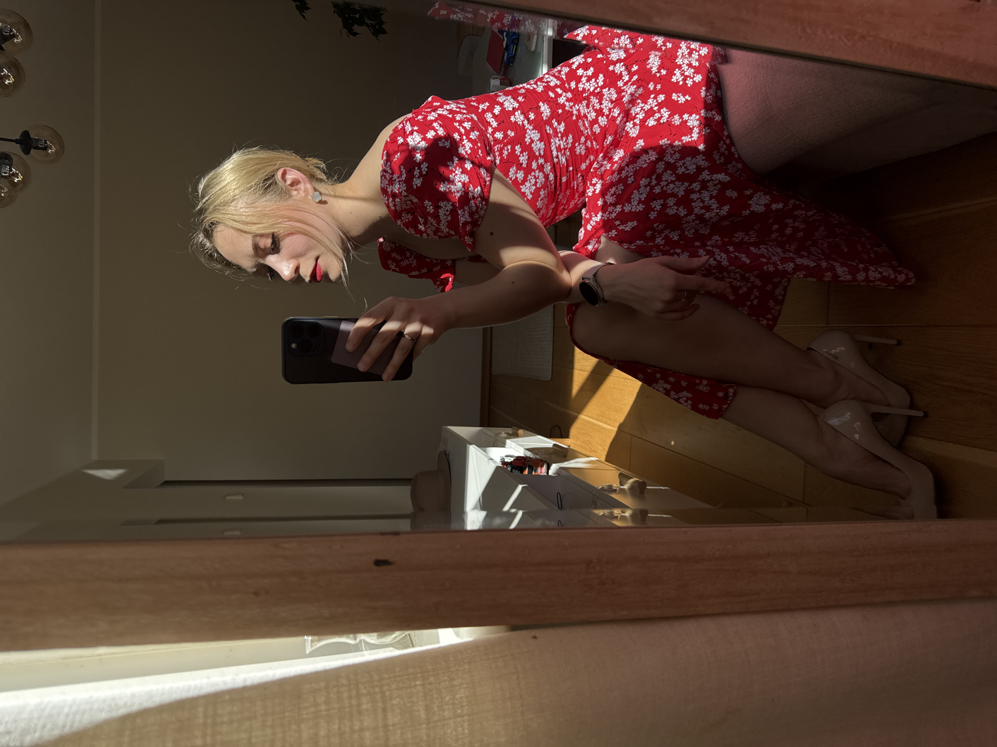 A woman is taking a selfie in a mirror. She is wearing a red and white floral dress and nude heels. The location appears to be an interior setting with a wooden floor and furniture. Without additional information or context, it is impossible to determine if the location is in Riga, Latvia.