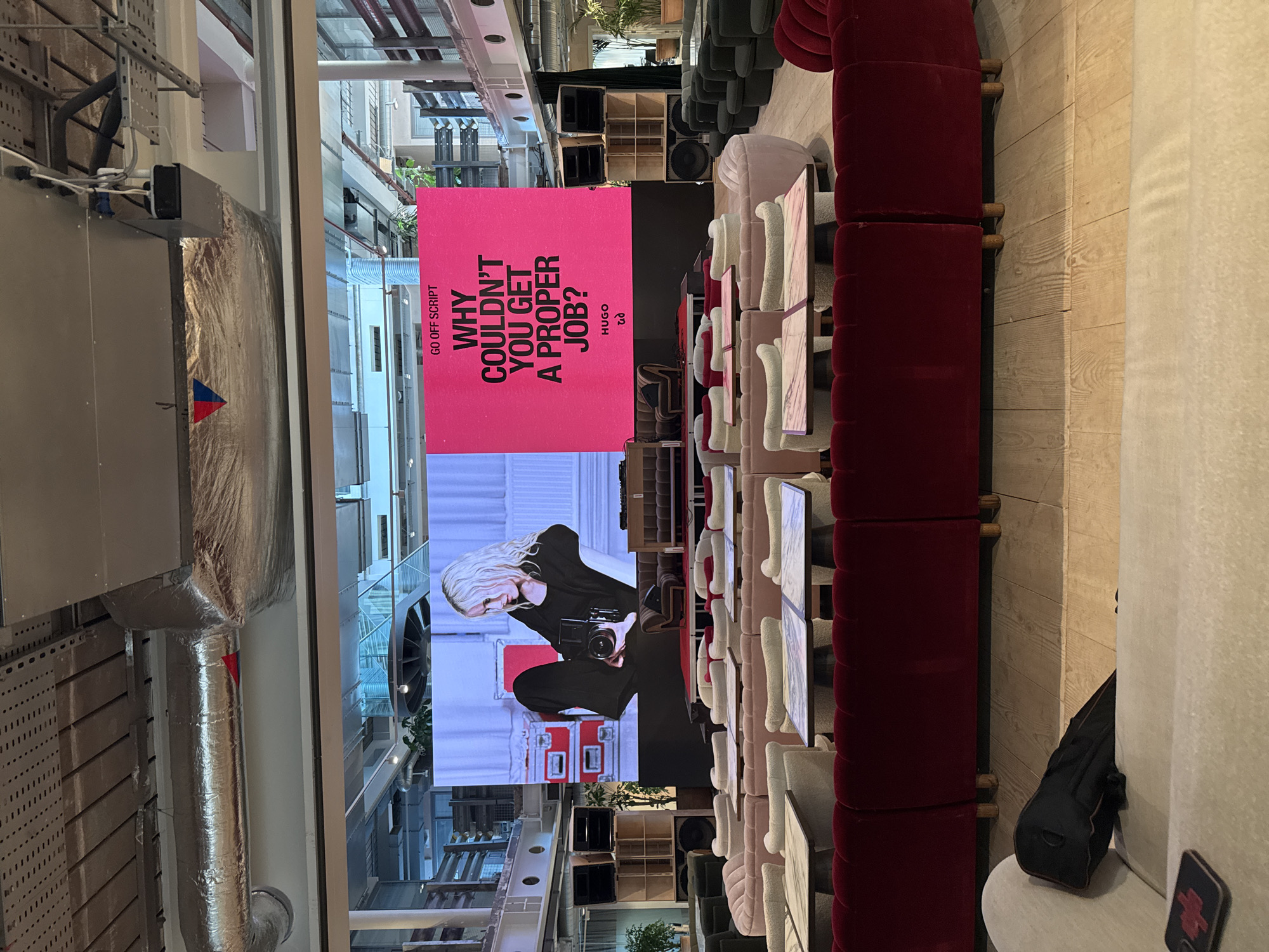 The image shows the interior of a large space, likely a store or event venue in London, United Kingdom. A large screen displays an advertisement with the text "GO OFF-SCRIPT. WHY COULDN'T YOU GET A PROPER JOB? HUGO." Below the text is an image of a photographer with a camera. The space is furnished with rows of tables and chairs, upholstered in varying shades of pink and red. The walls are white. There are ductwork and mechanical systems visible on the ceiling. Further in the background, there are open spaces with greenery. In the foreground is a black bag placed on a chair or couch.