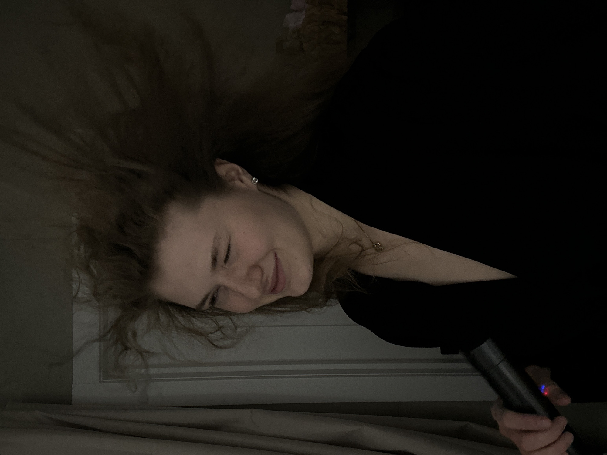 A woman is lying down, holding a black cylindrical object that appears to be a microphone. The woman has fair skin, brown hair, and is wearing a black top. She is smiling with her eyes closed. The background consists of a dimly lit room with a window frame visible. No specific details indicate the location being in Kazan, Russia.