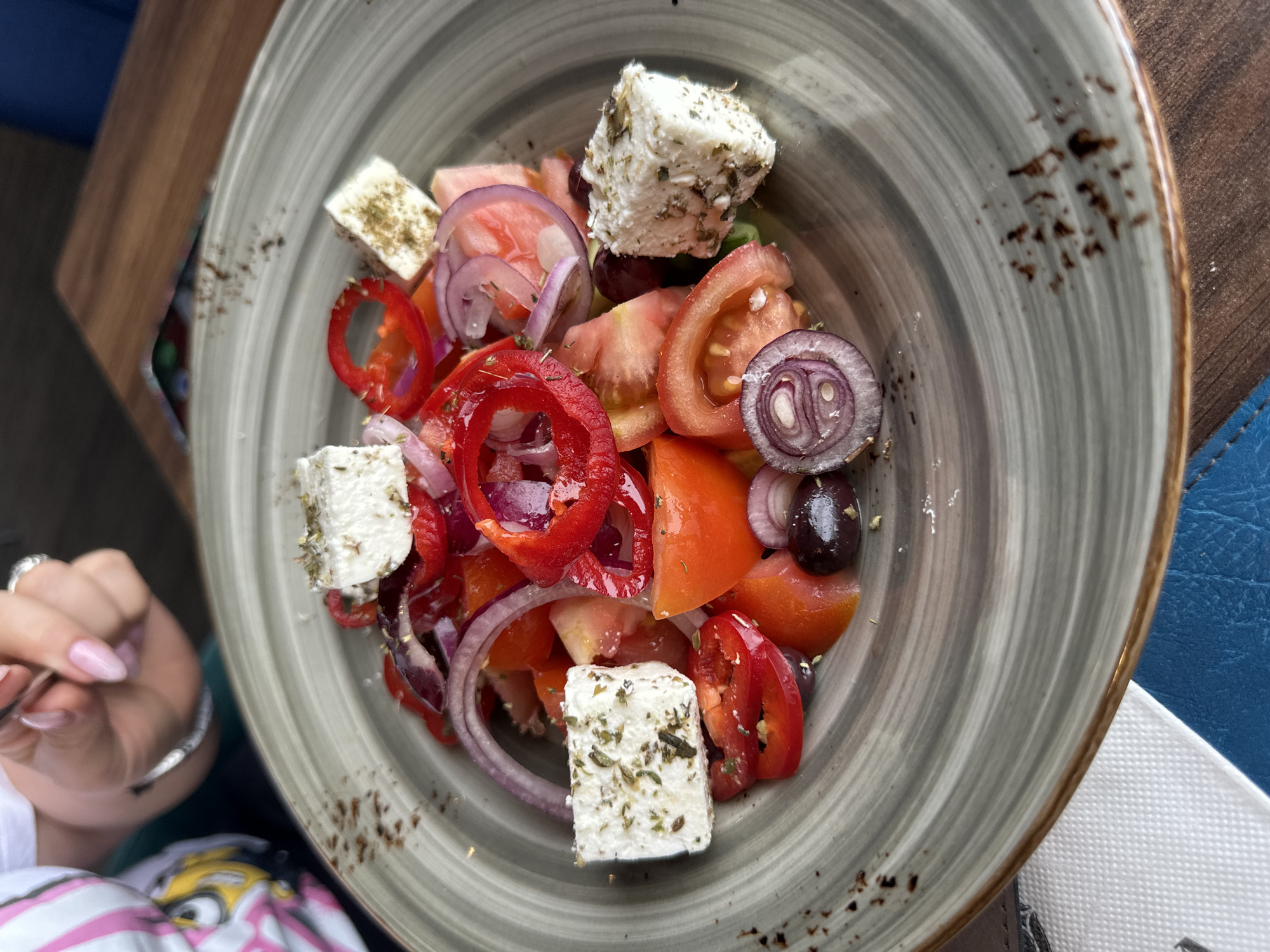The image shows a plate of Greek salad. The salad contains tomatoes, red onion, red bell pepper, olives, and feta cheese cubes. The feta is seasoned with herbs. The plate is grey with a brown rim. A hand holding a fork is visible to the left of the plate. The salad is on a wooden table, and a blue object is seen on the right side of the table. The location is Vidin, Bulgaria.