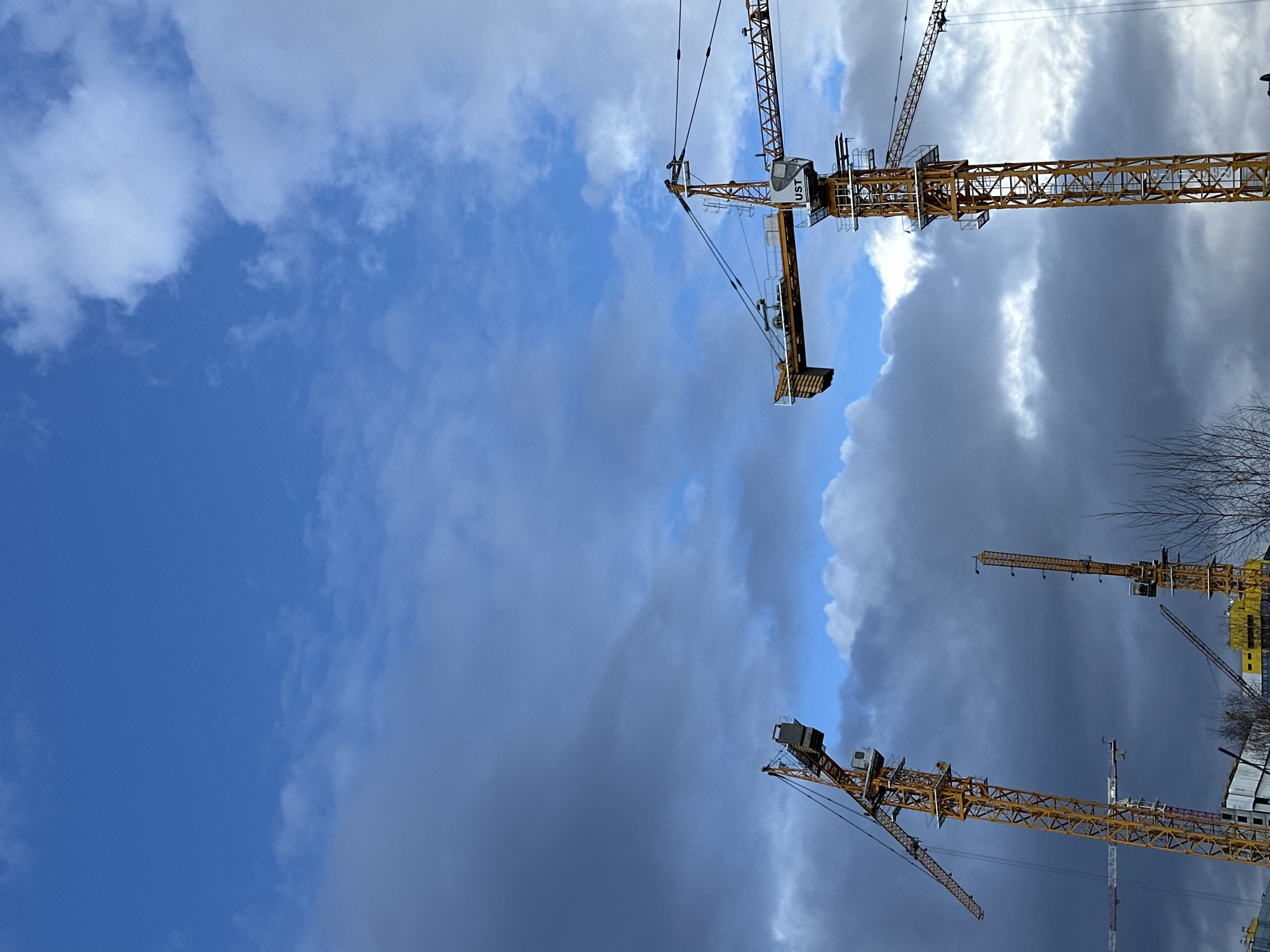 The image displays a construction site dominated by multiple large, yellow tower cranes set against a dramatic and varied sky.
**Subjects:** The primary subjects are several industrial construction cranes, characterized by their lattice-like yellow and orange structures. One prominent crane, located in the upper right quadrant, features a cabin with visible text. Another crane is partially visible in the lower right, and elements of what appear to be other cranes or parts of the construction site are scattered throughout. The sky fills a significant portion of the frame, showcasing a mix of clear blue, fluffy white, and dark, heavy grey clouds. Partially constructed building structures are faintly visible towards the lower right edge of the frame, blending with the darker clouds. A few bare tree branches are also discernible on the far right.
**Activity or event taking place:** The presence of multiple cranes indicates ongoing construction or development, although no active work or personnel are visible in the frame.
**Notable details:** The time of day appears to be daytime, with natural light illuminating the scene. The weather is dynamic, characterized by a striking contrast between bright blue sky and white clouds on the left and upper parts, transitioning into much darker, heavy grey clouds on the right and lower sections. This suggests a rapidly changing weather pattern, possibly before or after a storm, or a mix of sunny and overcast conditions.
The image is oriented vertically, creating an interesting perspective looking upwards towards the cranes and sky.
The provided location context is Yekaterinburg, Russia, though there are no explicit visual cues within the image itself to confirm this.
**Visible text:** The letters "UST" are clearly visible on the cabin of the most prominent crane in the upper right.
