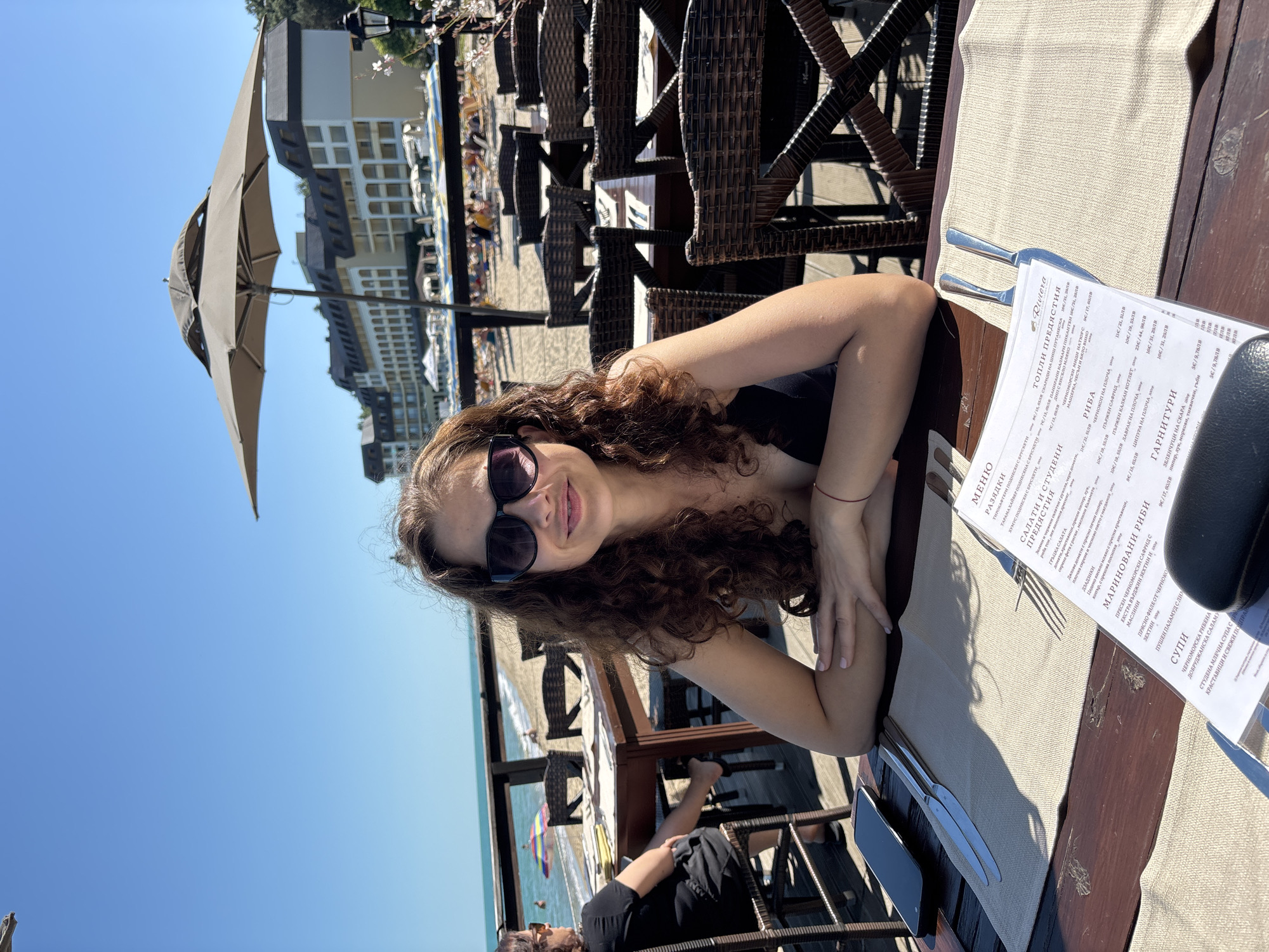 A woman with curly brown hair and sunglasses is sitting at a table on a terrace overlooking a beach and the Black Sea. She is wearing a black swimsuit top and has her arms crossed on the table. A menu, cutlery, and a smartphone are on the table. In the background, there are other tables and chairs, beach umbrellas, and a hotel building. The scene is set in Varna, Bulgaria, likely at a restaurant on the Varna beach promenade. The weather is sunny and clear.