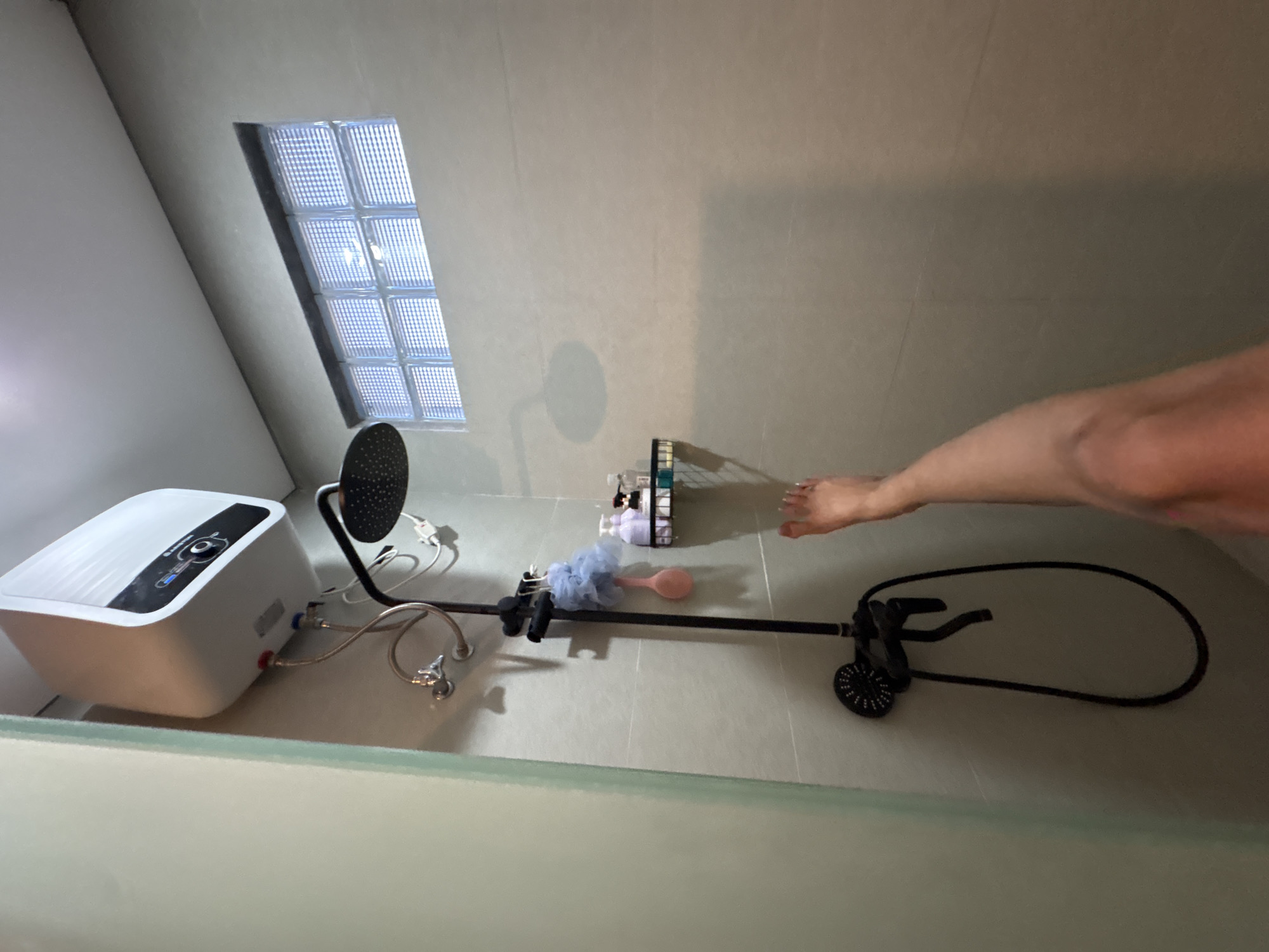 The image depicts a bathroom interior, possibly in Unknown, Unknown. The room is tiled in a neutral tone, with a small glass block window providing natural light. A white water heater is mounted on the wall, connected to a shower system with a black shower head. A black grab bar is positioned on the wall, along with an open bottle rack and a loofah, indicating the presence of personal hygiene products. A foot and part of an arm are visible near the grab bar, suggesting someone is present in the bathroom. The overall impression is of a functional and well-maintained bathroom space.