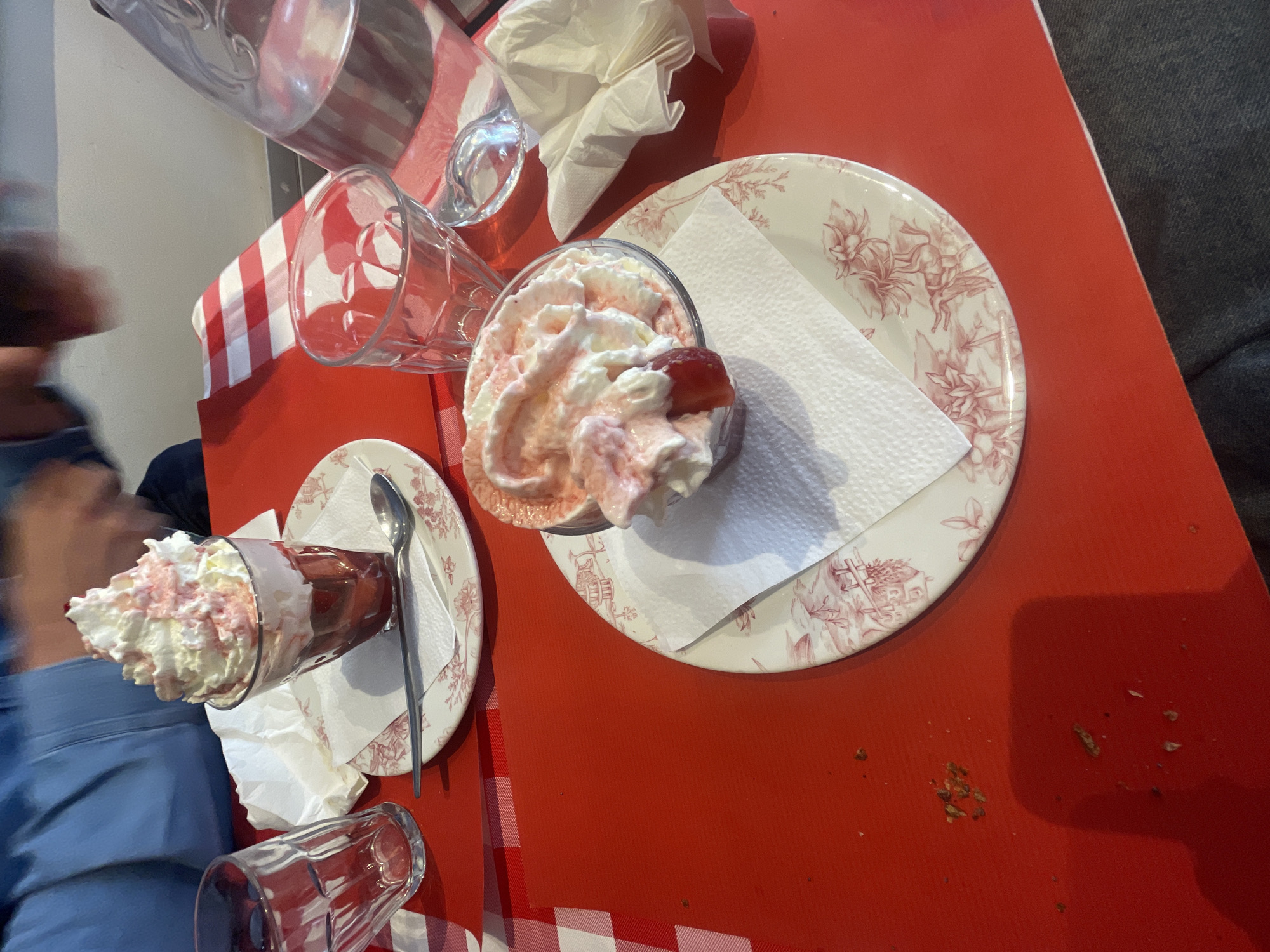 A high-angle shot showcases a red and white checkered tablecloth setting in what appears to be a restaurant, possibly in Paris, France. Two glasses of strawberry sundae, topped with whipped cream, are arranged on decorative plates with matching floral patterns. A single spoon rests on the plate beside one sundae. Additional glasses, some empty and one filled, are also visible on the table, along with crumpled napkins. Traces of crumbs are scattered on the red portion of the tablecloth. A person dressed in a blue garment is partially visible on the left side of the image, while a dark-clothed individual is situated on the right. The arrangement suggests a dining experience.