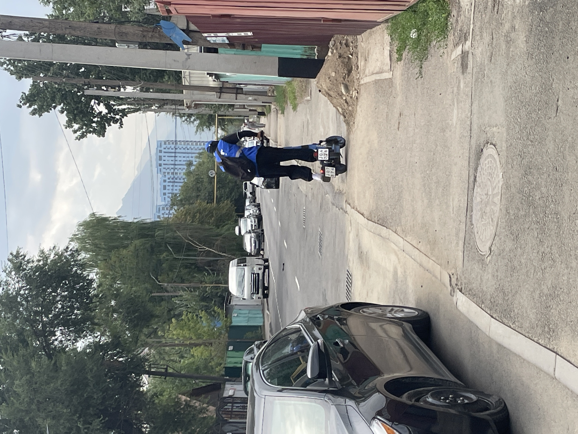 The image shows a man riding an electric scooter on a sidewalk in Almaty, Kazakhstan. The man is wearing a blue and black outfit and a backpack. The scooter has a license plate that reads "JD4 726". In the background, there are cars on the street, trees, buildings, and mountains. The weather appears to be overcast.