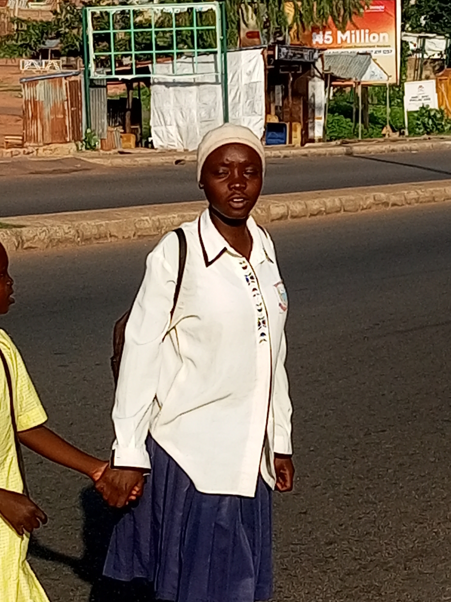 In Garko, Nigeria, two individuals, one an adult and the other a child, walk hand-in-hand along a paved road. The adult, wearing a white shirt and a navy blue skirt, has a backpack on their left shoulder and is looking towards the camera. The child, dressed in a yellow outfit, is walking alongside the adult. In the background, a street scene unfolds with various structures, including corrugated metal shacks and a green-gridded structure. A large advertisement board displaying "N5 Million" is visible, suggesting commercial activity in the area. The road surface is asphalt, bordered by a stone curb.