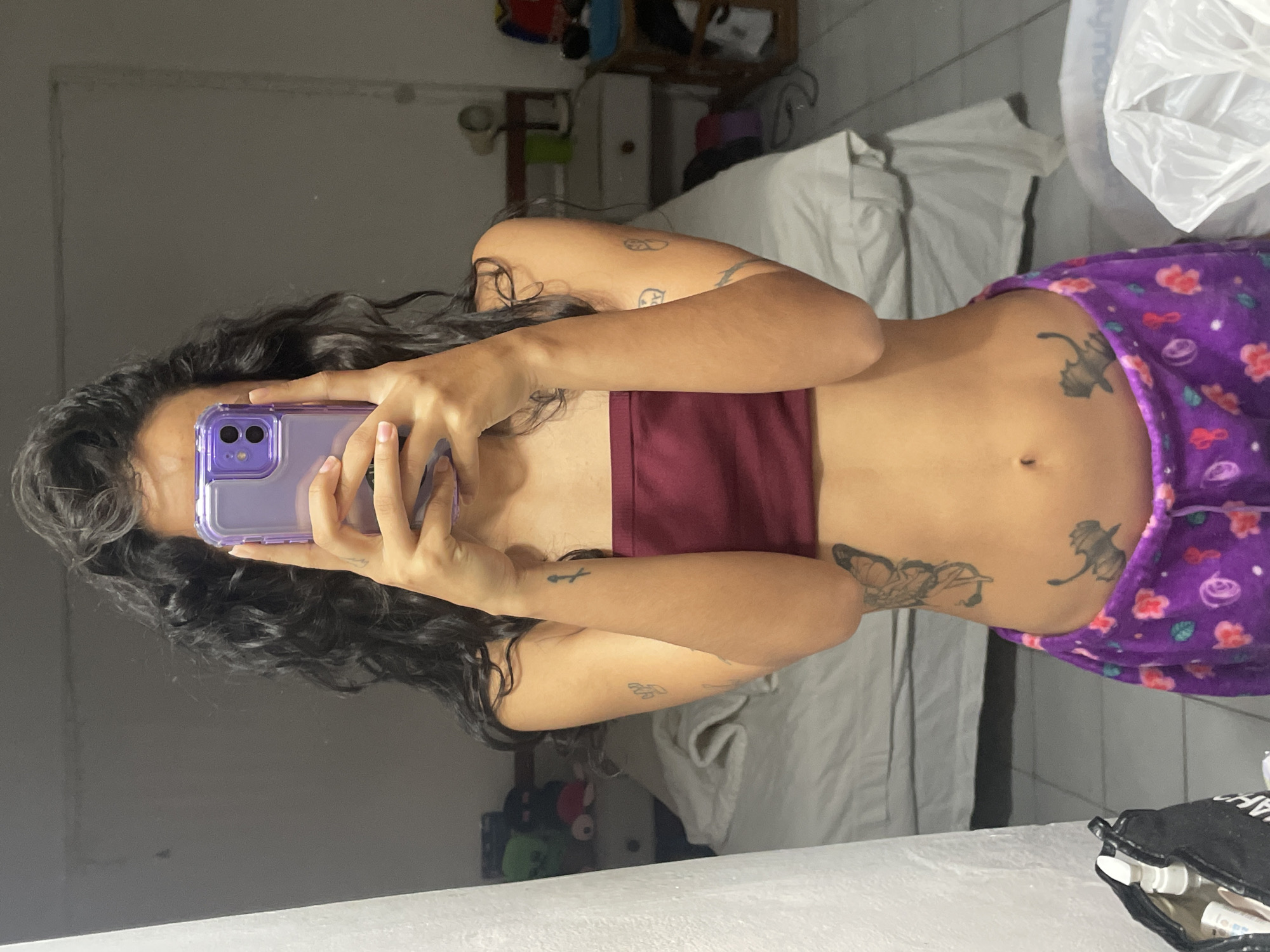 A person is standing in front of a mirror, holding a purple phone to take a picture of their reflection. The person has dark, curly hair and is wearing a maroon strapless top and purple pajama bottoms with a floral print. They have tattoos on their arms, stomach, and hip, including a butterfly and what appears to be a cat-like creature. The background shows a bedroom with a bed, a nightstand, and a closet. The room appears to be illuminated by artificial light. The image is a full-body selfie taken indoors, likely in a residential setting. There is no indication of the city of Porlamar, Venezuela, in the image itself.