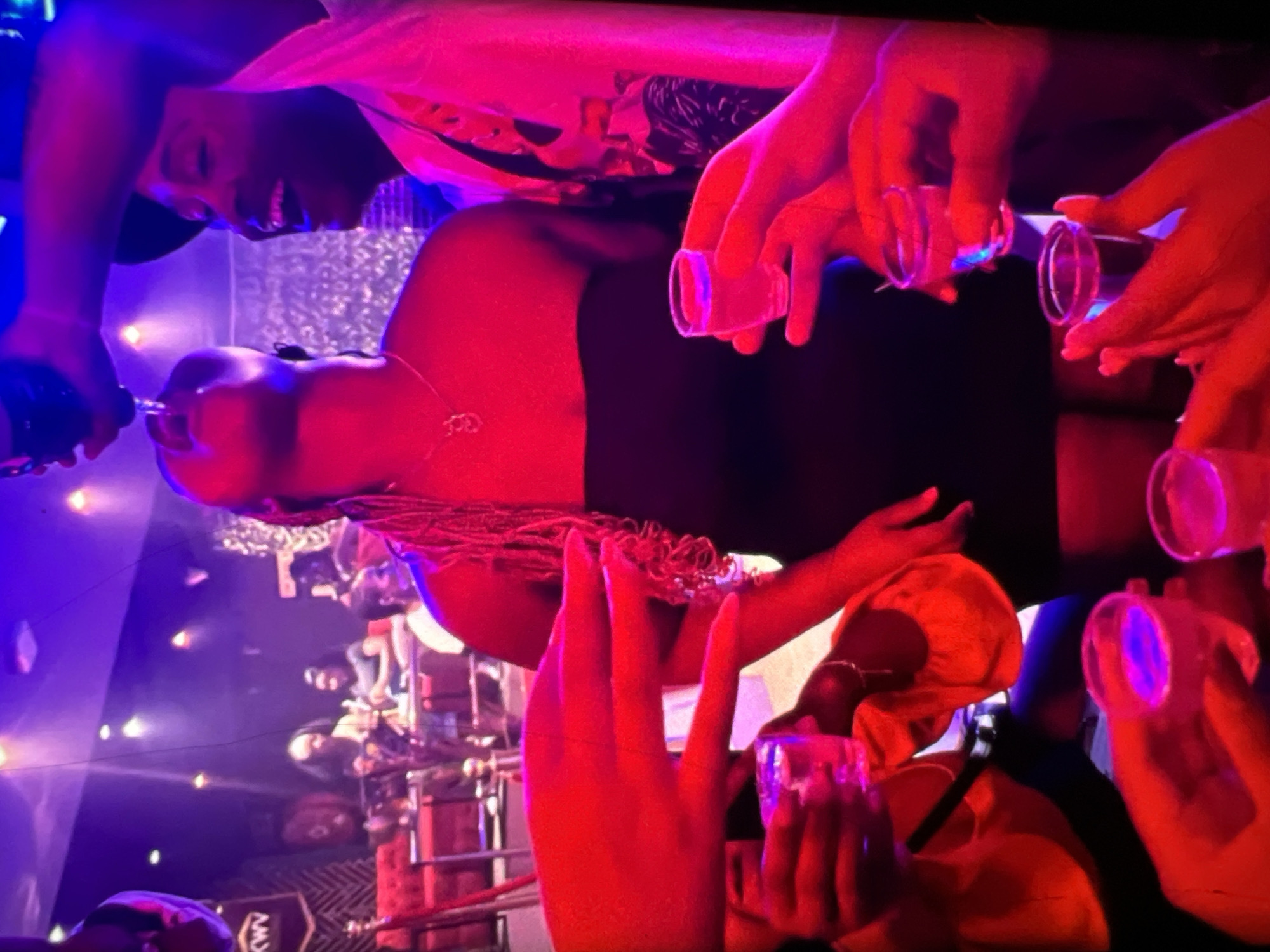 The image depicts a group of individuals in what appears to be a dimly lit, indoor venue, possibly a nightclub or bar, illuminated by red and purple lighting. In the foreground, several hands are visible, holding small shot glasses filled with a clear liquid. The primary focus is a woman in a black strapless dress who is tilting her head back, with a shot glass being offered to her mouth from above. A man to her right, wearing a white shirt and a dark hat, is smiling and looking towards her. The background shows other patrons and seating areas. The overall scene suggests a celebratory or social gathering where drinks are being consumed. The presence of the "KWV" logo on a banner in the background suggests a possible connection to a South African wine and spirits producer, which has distribution in Lagos, Nigeria.