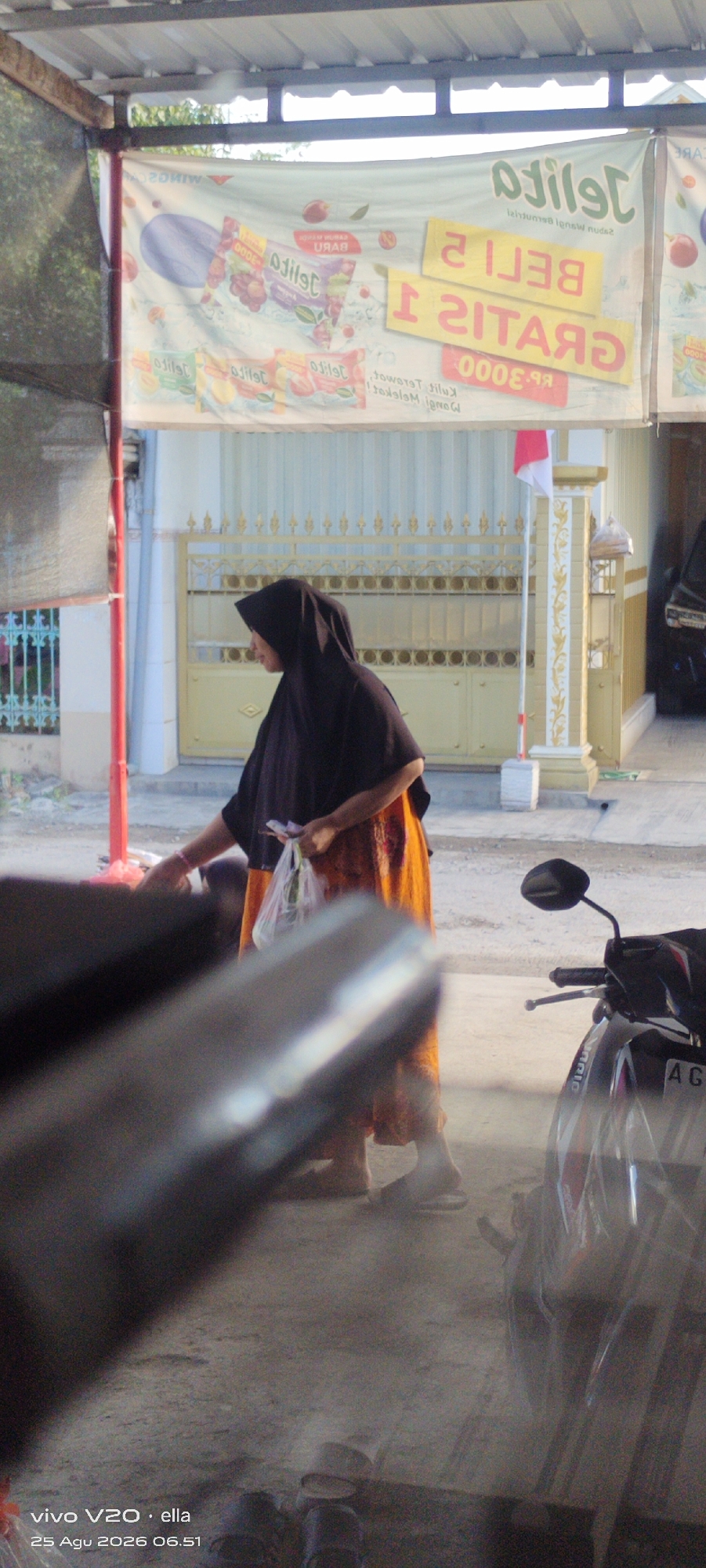 A woman wearing a black hijab and an orange skirt is standing near a red pole and a gate. She is holding a plastic bag in her right hand. In the background, a banner with Indonesian text and images of fruit is visible. The scene appears to be outdoors, with a concrete ground. A motorcycle is parked to the right of the woman. The location is identified as Simo, Indonesia.