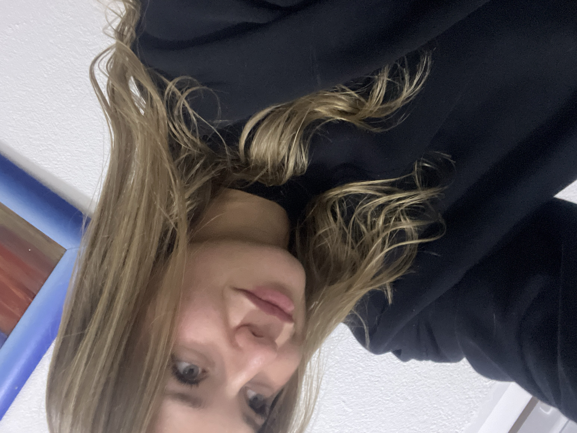 The rotated image displays a young woman in a close-up, selfie-style portrait. She has long, wavy blonde or light brown hair that frames her face and spills over her shoulders. She is wearing a dark, possibly black, hooded top. Her head is tilted, and she looks towards the viewer with a soft, relaxed expression, featuring a slight smile and visible eyes. Her lips are full.
The background is predominantly a textured, light-colored surface, appearing to be an indoor wall or ceiling. To the upper left side of the frame (as depicted in the rotated image), a blurry, colorful object with blue and red sections is partially visible, hinting at furniture, artwork, or an inflatable structure in the background. The lighting suggests an indoor setting, and no specific time of day or weather conditions can be discerned. There is no visible text. The location context for this image is Sotonići, Montenegro.