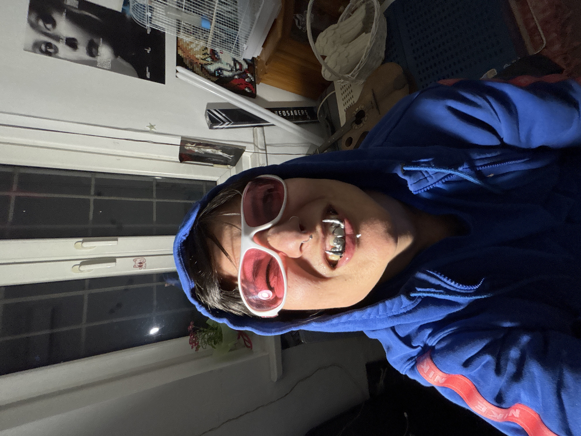 A person wearing a blue hooded Nike sweatshirt and pink sunglasses is angled towards the camera. They have braces and a nose ring. Behind them, a window with plants on the sill is visible. On the wall, there's a black-and-white poster, a fan, and a sign that reads "NEOSABERS." A guitar and a white basket are also visible. There is no information available about the city Sofia, Bulgaria.