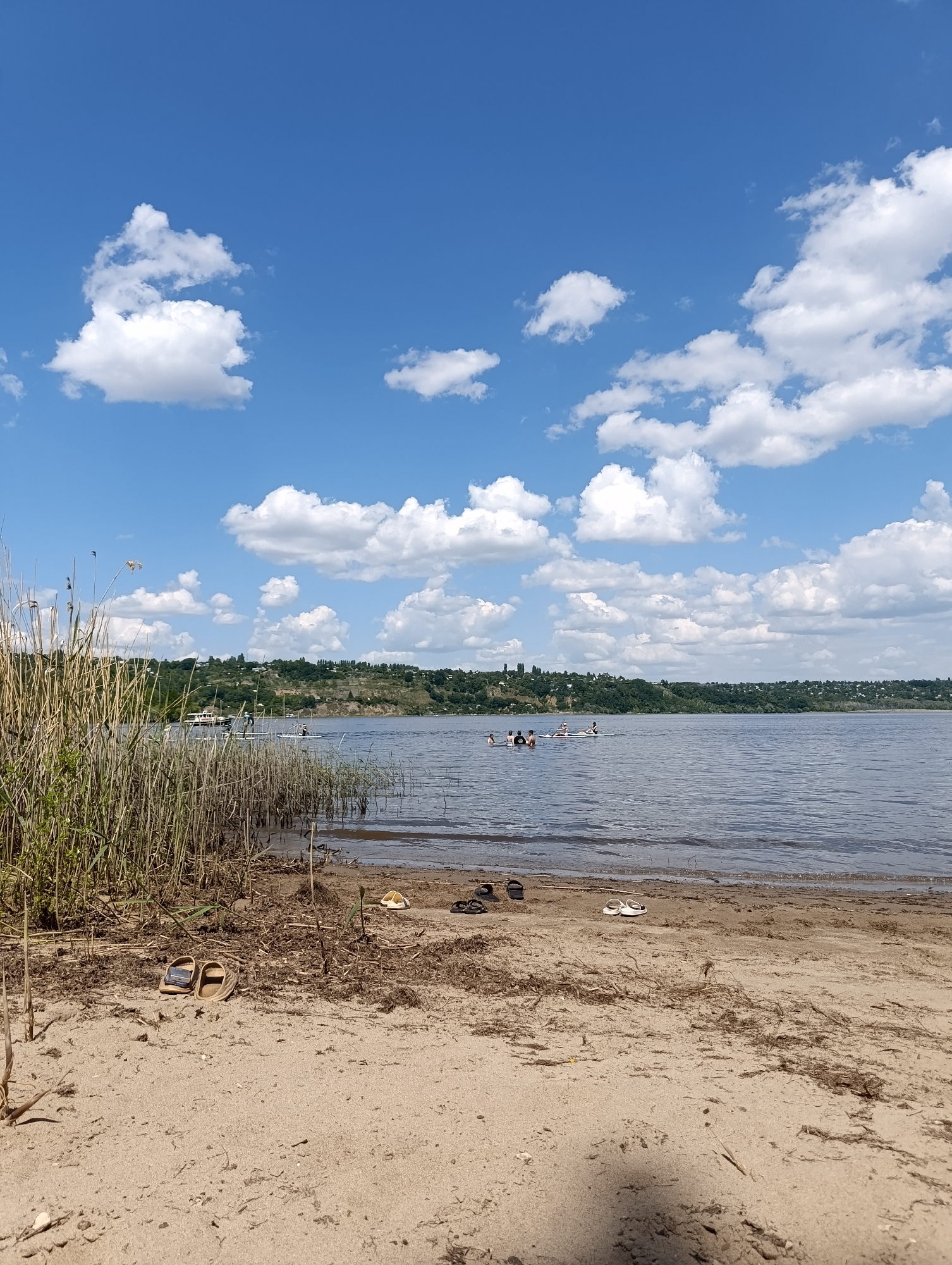 This image depicts a serene summer day at a river beach. In the foreground, a sandy shore is adorned with dry reeds and scattered debris. A pair of flip-flops sits near the water's edge. Beyond the beach, the calm river stretches out, with several people visible in the water and on paddleboards. A boat can also be seen anchored on the water. The background features a lush green hillside dotted with houses, under a bright blue sky filled with fluffy white clouds. This scene is characteristic of the Volga River embankment in Saratov, Russia.