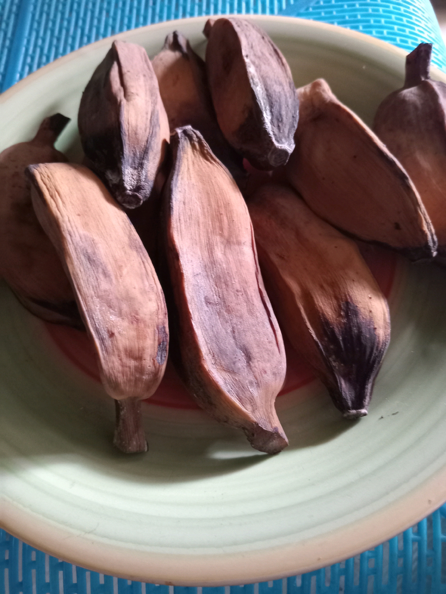 The image shows a close-up, overhead view of a light green ceramic plate filled with dried, peeled bananas. The bananas are various shades of brown, with some exhibiting black spots indicating ripeness or age. They are arranged in a somewhat disordered pile on the plate, with their stems and blossom ends visible. The plate has a light brown rim and sits on a surface that appears to be a blue, textured mat or placemat. The lighting suggests an indoor setting, possibly in Randuagung, Indonesia, given the context of the request, although no definitive environmental cues like architecture or specific Indonesian flora are visible. There are no people or animals present in the image.