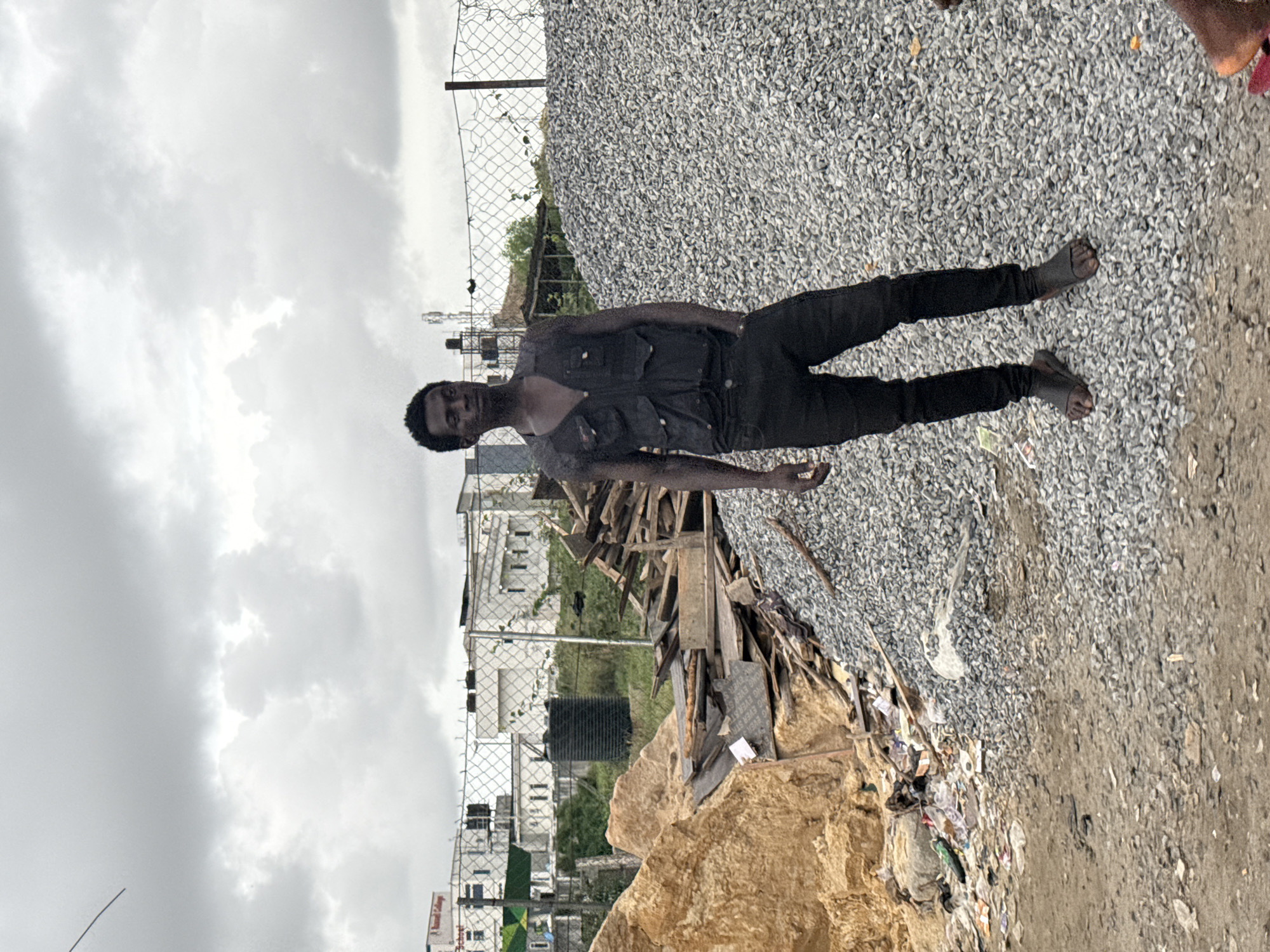 A medium-shot captures a dark-skinned man wearing dark clothes and sandals lying at an oblique angle on a gravel surface and next to a pile of garbage and discarded wood. Behind him, there is a wire fence and buildings, possibly in Lekki, Nigeria. The sky is overcast. The scene appears to be outdoors.