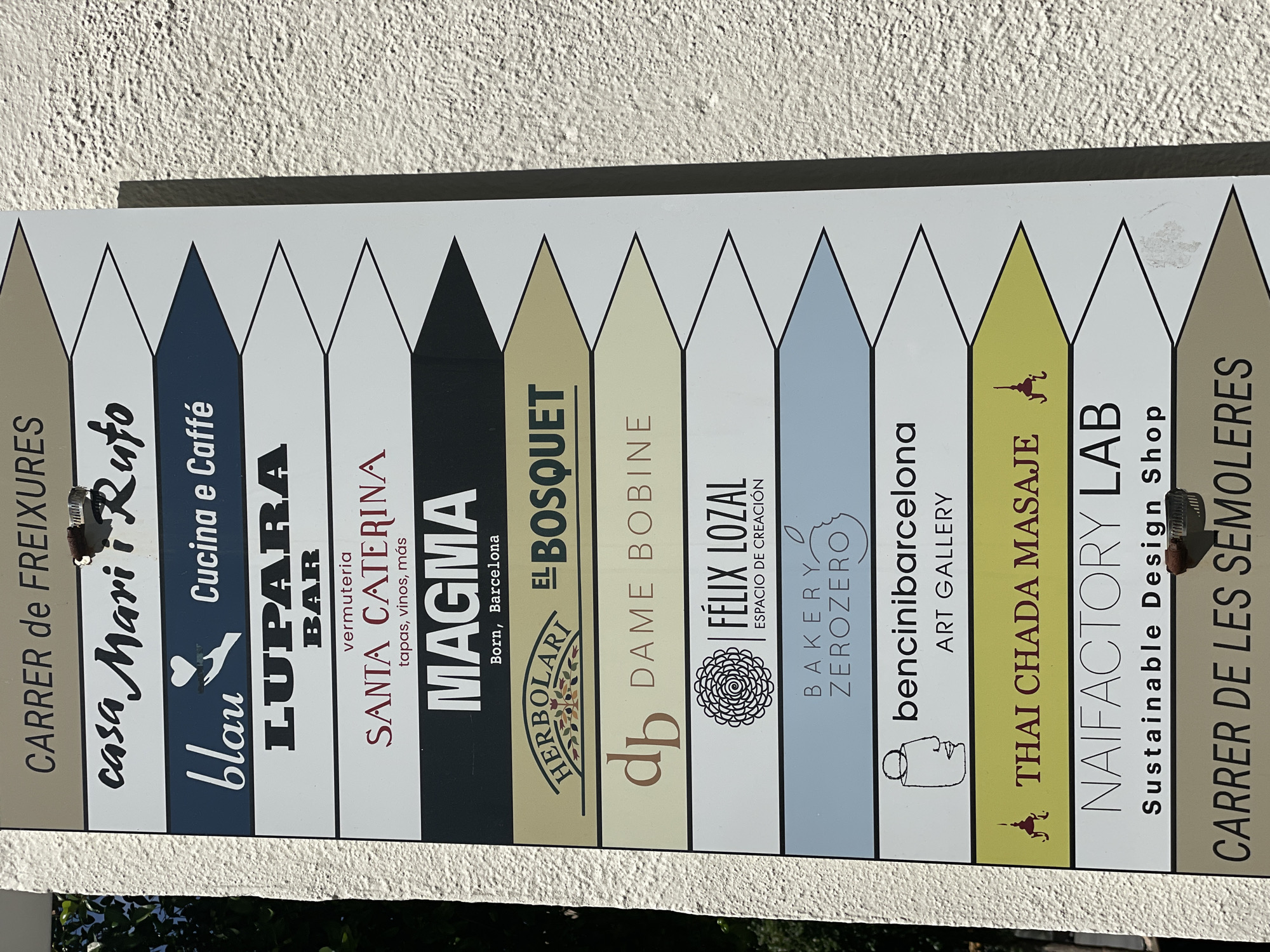 A vertically oriented commercial directional sign, affixed to a textured, off-white building wall, is depicted under bright sunlight. The sign features multiple horizontally aligned, arrow-shaped panels pointing to the right, listing various businesses and two street names in Barcelona, Spain. From top to bottom, the sign displays: 1. A brown-gold arrow with "CARRER de FREIXURES" in white capital letters. 2. A white arrow with "casa Mari i Rufo" in black cursive font. A small, dark, clip-like object is attached to the upper right edge of this panel. 3. A dark blue arrow with "blau" in white cursive, an icon of a white hand holding a heart, and "Cucina e Caffé" in white. 4. A white arrow with "LUPARA" in large, bold black capital letters and "BAR" in smaller black capital letters below it. 5. A white arrow with "vermuteria" in small red text, "SANTA CATERINA" in a larger red serif font, and "tapas, vinos, más" in small red text. 6. A black arrow with "MAGMA" in large white sans-serif font and "Born, Barcelona" in smaller white text, confirming the location. 7. A tan/beige arrow with "HERBOLARI" in an arched brown font above a brown olive branch-like logo, and "EL BOSQUET" in bold brown text. 8. A light beige/cream arrow with a stylized "db" in brown script and "DAME BOBINE" in brown text. 9. A white arrow featuring a black circular, spiral flower-like logo, "FÉLIX LOZAL" in black sans-serif font, and "ESPACIO DE CREACIÓN" in smaller black text. 10. A light blue arrow with "BAKERY" and "ZEROZERO" in black text, accompanied by an outline of an apple or similar fruit. 11. A white arrow with a black outline drawing of a stylized human head/bust, "bencinibarcelona" in black, and "ART GALLERY" in black. 12. A yellow-green arrow with "THAI CHADA MASAJE" in red text and a small red stylized figure or symbol. 13. A white arrow with "NAIFACTORY LAB" in black sans-serif font and "Sustainable Design Shop" in smaller black text. Another small, dark, clip-like object is attached to the right side of this panel, and a light grey smudge is visible below it. 14. A bottom brown-gold arrow with "CARRER DE LES SEMOLERES" in white capital letters. The building wall exhibits a coarse, stucco-like texture. A clear shadow is cast to the right of the sign, indicative of direct sunlight from the left. In the upper left background, a patch of