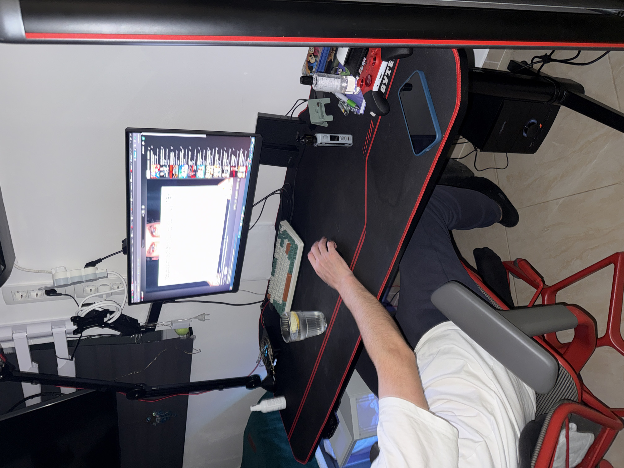 The image shows a person sitting at a desk with a computer monitor, keyboard, mouse, phone, and a glass of water with a lemon slice. The desk has a black surface with red trim. A game controller and other small items are on the desk. The person is wearing a white shirt and dark pants. The chair is red and black. A speaker is visible under the desk. The wall behind the desk is white and has a power strip mounted on it. The environment appears to be an indoor space, possibly a home office.
