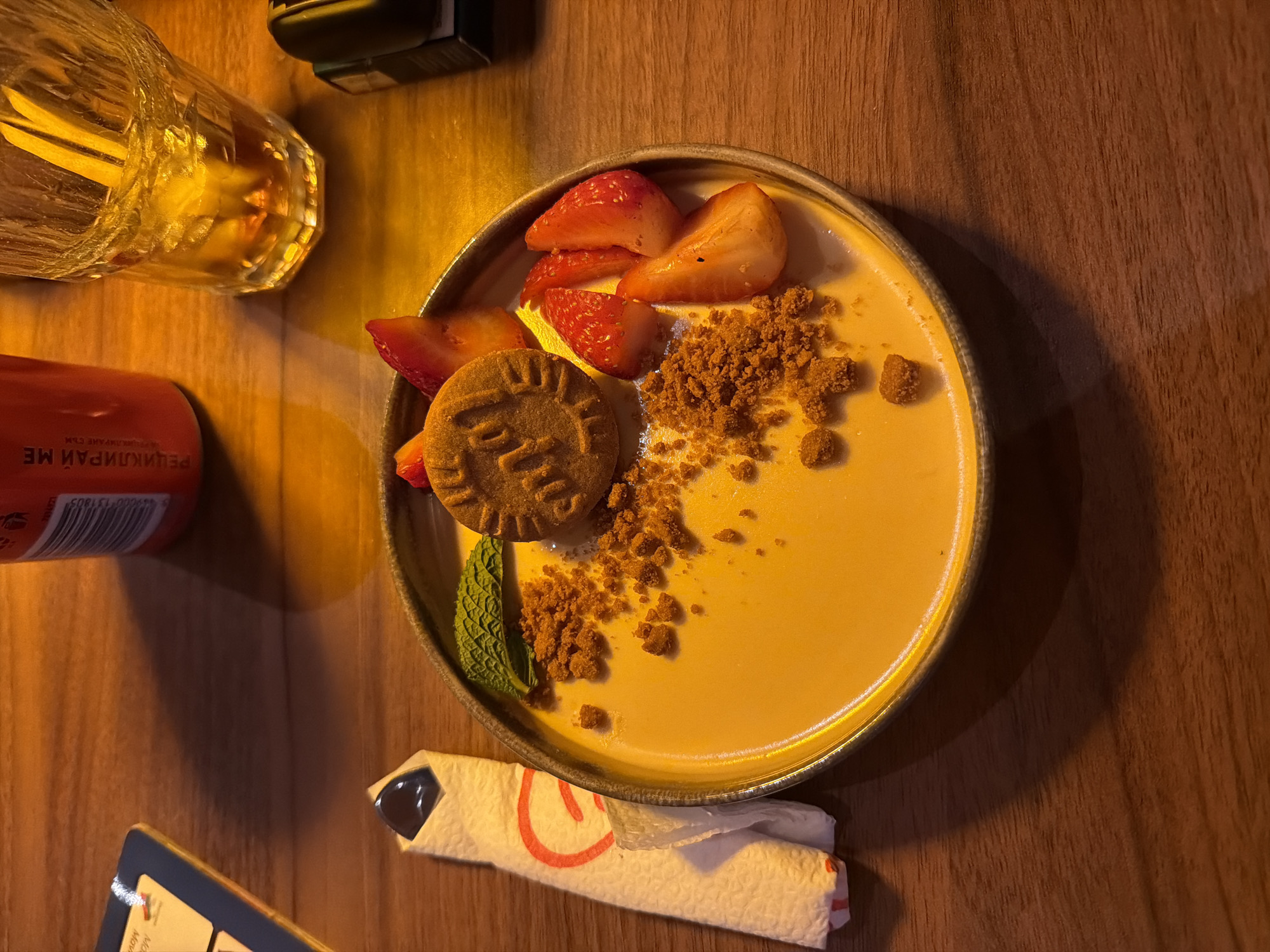 The image depicts a dessert served in Sofia, Bulgaria. The dessert consists of a light-colored custard or pudding base in a shallow, dark-rimmed dish. It's topped with sliced fresh strawberries, crumbled biscuit pieces, and a small sprig of mint. A whole biscuit is also placed on the side. To the left of the dish is a partially visible glass of an amber-colored drink (possibly whiskey), a can of soda (red), and a small black box, possibly containing sweeteners. A wrapped napkin with a logo (partially visible) is placed below the dish. The background is a wooden table. The lighting is warm, suggesting indoor illumination.